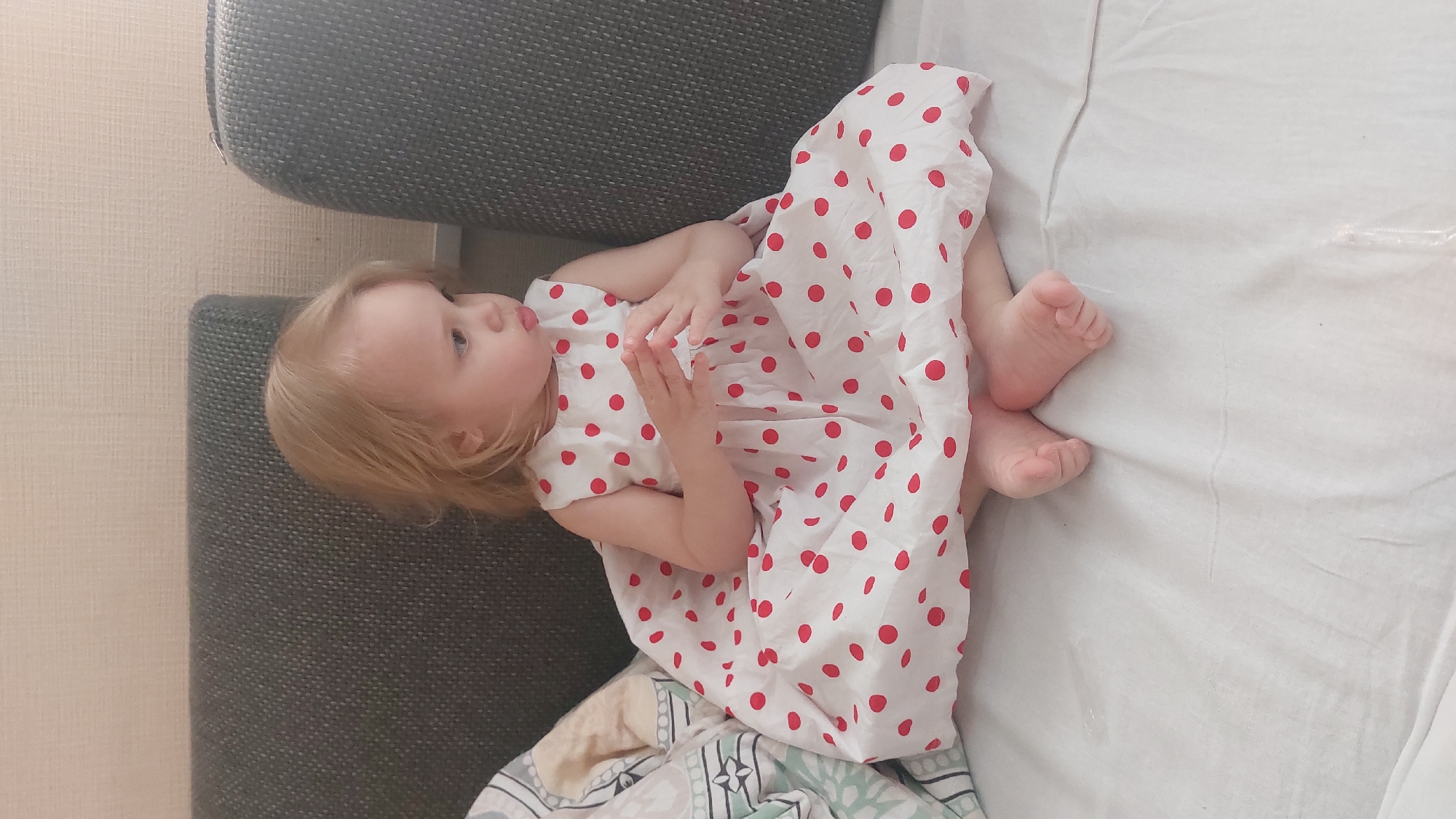 A toddler with blonde hair is lying on a couch and bed, in the city of Novosibirsk, Russia. The toddler is wearing a white dress with red polka dots. The couch is grey and the bed has white sheets. The toddler's feet are visible on the bed.