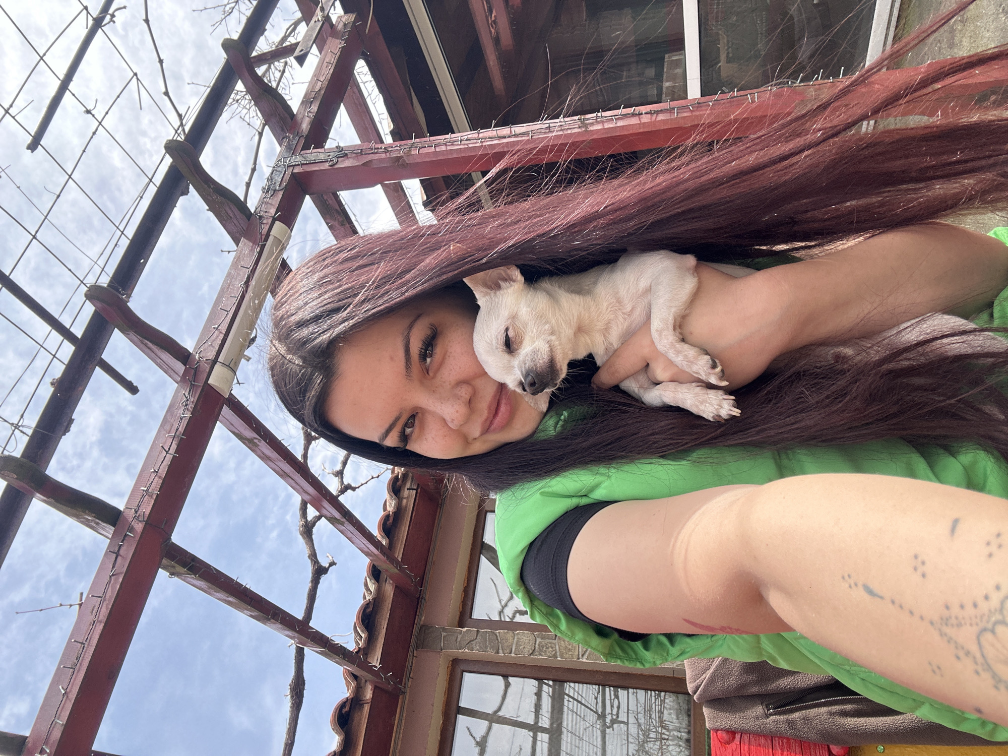 A woman holds a small, sleeping white dog in her arms. She is wearing a green jacket over a black top. Her long, dark hair partially obscures the dog. The background includes a weathered wooden pergola, a building with windows, and a partially visible cloudy sky. The location is Chubra, Bulgaria, as indicated in the image metadata.