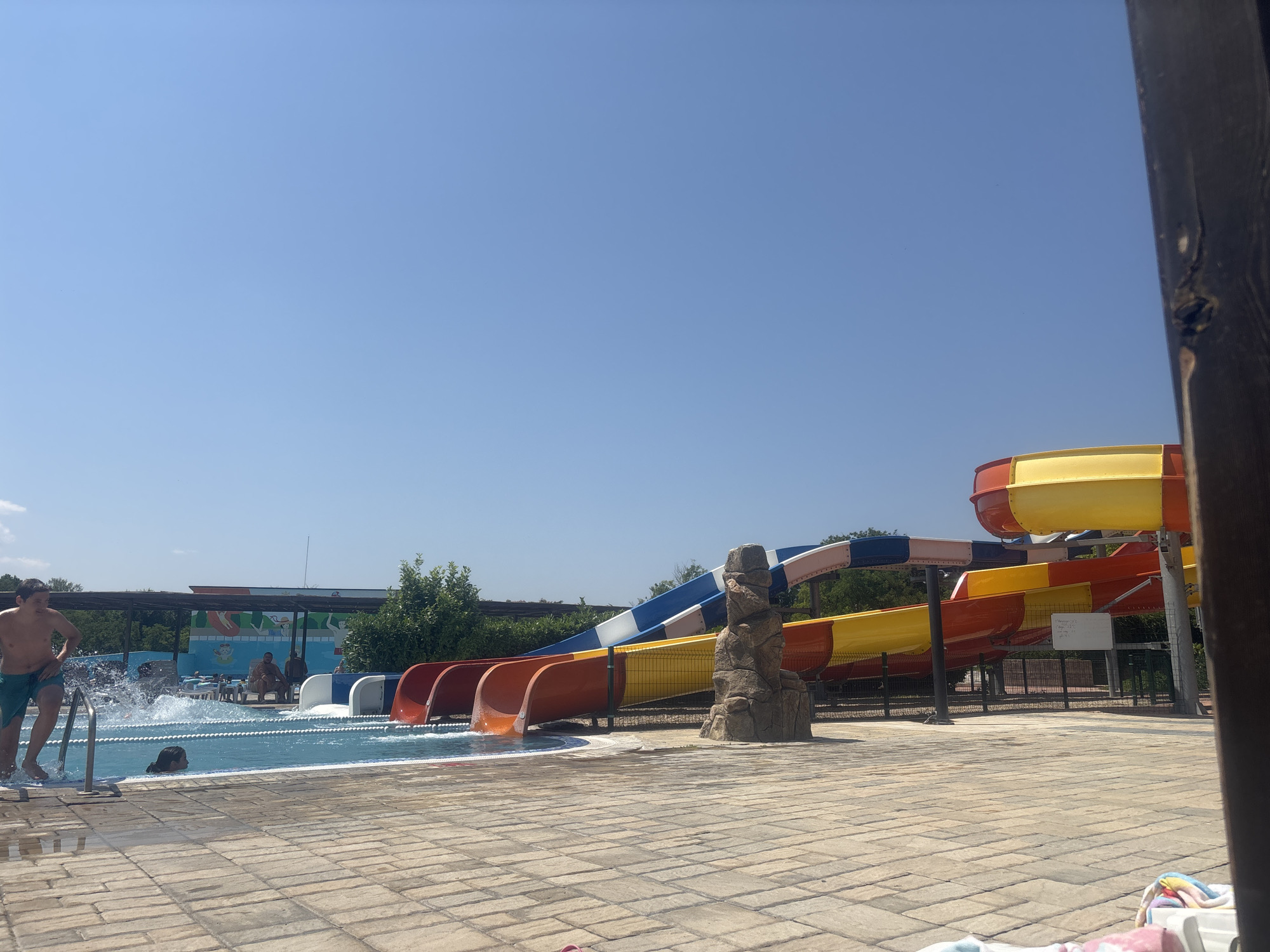 The image depicts a sunny day at a water park, likely located in or near Pobeda, Bulgaria. In the foreground, a tiled patio area is visible, with a portion of a wooden post on the right. To the left, a swimming pool with clear blue water is present, with a man in green swim trunks entering the water, causing a splash. A child is swimming in the pool. Several red and orange water slides are prominently featured in the midground, with a blue and white slide above them. These slides appear to be made of brightly colored plastic. In the background, there is lush green foliage and trees, as well as a building with a mural depicting a water park scene. The sky is a clear, bright blue, indicating fair weather. A few other individuals are seen in the background, some sitting near the pool and others near the slides. The overall scene suggests a recreational environment for summer activities.