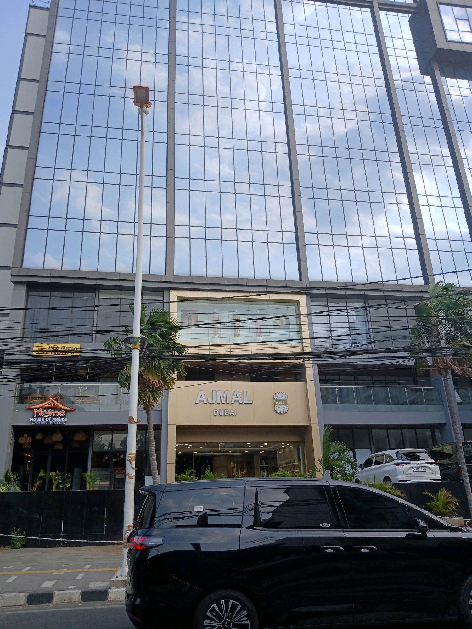 The image depicts the exterior of a modern building in Central Jakarta, Indonesia. The building features a glass facade reflecting the sky. On the ground level, there are several storefronts. To the left, a restaurant called "Meimo House of Meimo Manado Restaurant & Bar" is visible with its logo. To the right of Meimo, there is a building with signage that reads "AJMAL DUBAI" and a crest logo. In front of the building, a black minivan is parked, and a white car is parked on an elevated area behind the minivan. Utility wires are strung across the lower portion of the building. A street with a sidewalk and road markings is in the foreground. A lamppost stands near the center of the image. Palm trees are also visible on the property. The lighting suggests it is daytime.