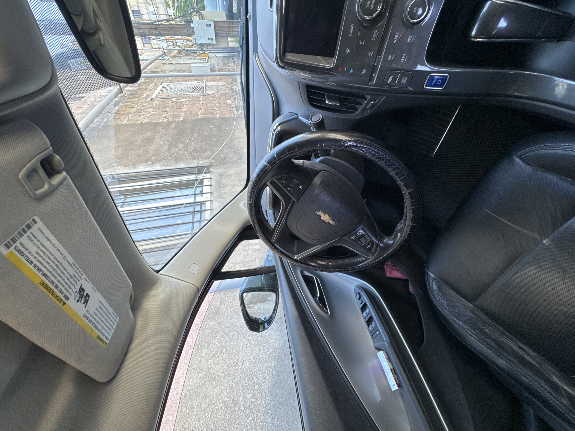 The image shows the interior of a car, likely a Chevrolet, with the steering wheel and dashboard prominently visible. Outside the car window, there is a view of an urban environment, with buildings and utility wires suggesting it could be in a city like Yerevan, Armenia. The view of the outside appears to be rotated clockwise. The sun visor is down, obscuring some of the view. The image focuses on the car's interior and the urban scene outside.