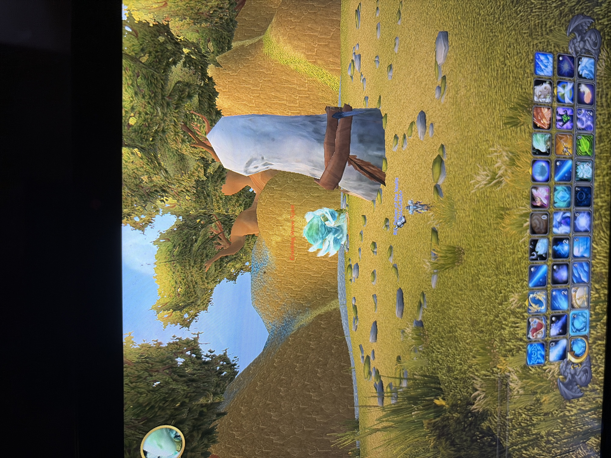 This is a screenshot of a video game, likely World of Warcraft, displaying a fantasy environment. The scene features rolling, yellow-tinted hills covered in sparse green grass and scattered rocks. Tall, leafy trees with brown branches frame the background. A prominent, light-colored, cylindrical stone monolith with a brown band around its middle stands in the foreground, slightly to the right of center. To the left of the monolith, a bright, crystalline, blue creature is visible. A small, humanoid character in armor is positioned in the middle ground, to the left of the monolith. The bottom of the screen shows a game interface with rows of icons representing abilities or items, and a small gargoyle-like figure on the left. The sky is a clear, bright blue. The overall impression is a serene, naturalistic fantasy landscape.