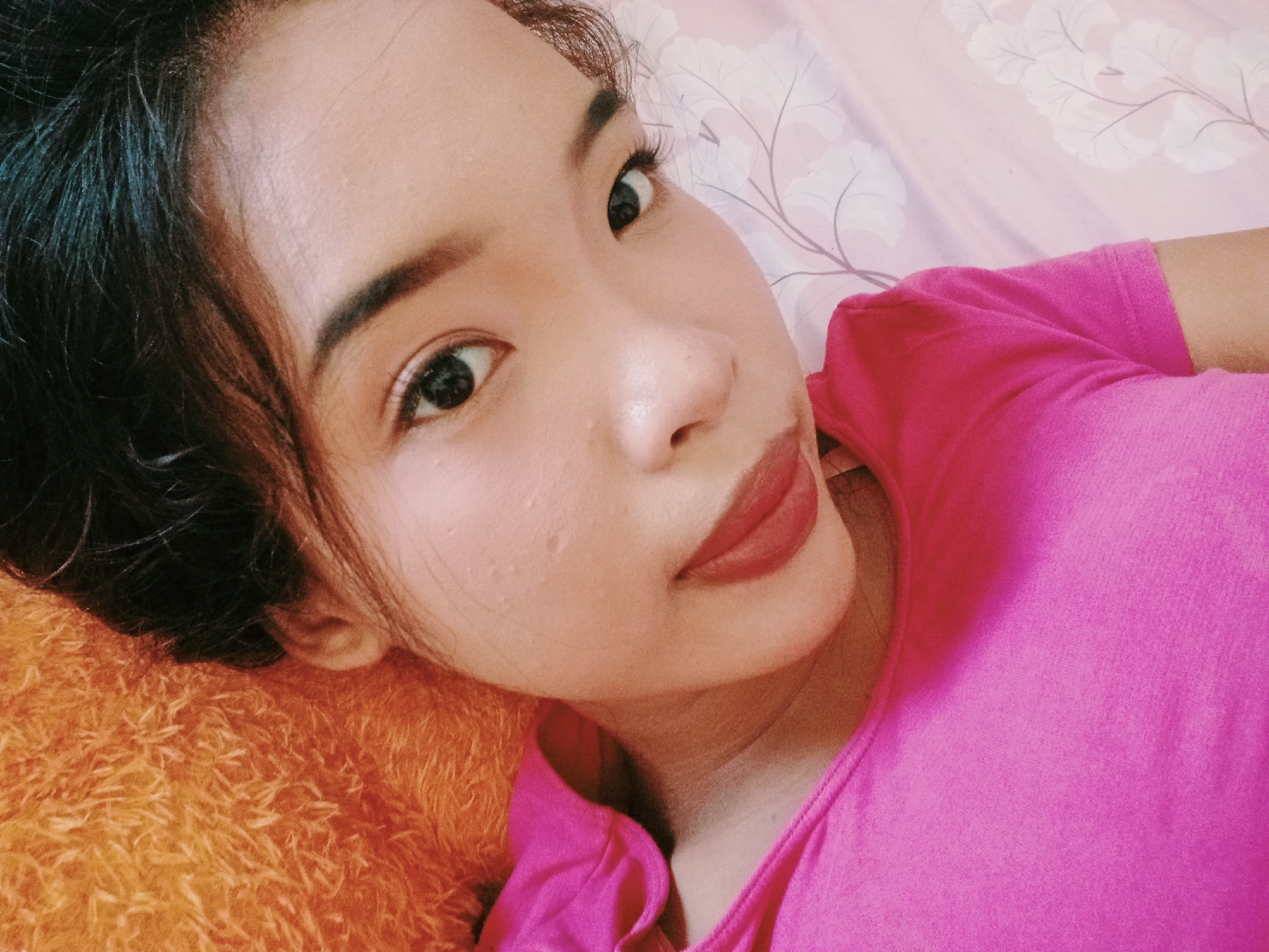 A close-up selfie of a young woman with dark hair and light brown eyes, wearing a bright pink t-shirt, is taken from a high angle. She is lying down on an orange, fuzzy surface on the left side of the image, and a pink sheet with a floral pattern is visible on the right. Her face is turned towards the camera, with her left eye and cheek more prominently featured. Her lips are painted a reddish-brown color. The lighting suggests it is daytime, possibly indoors, with a soft glow illuminating her face. The background is softly blurred, indicating a shallow depth of field, focusing attention on the subject.