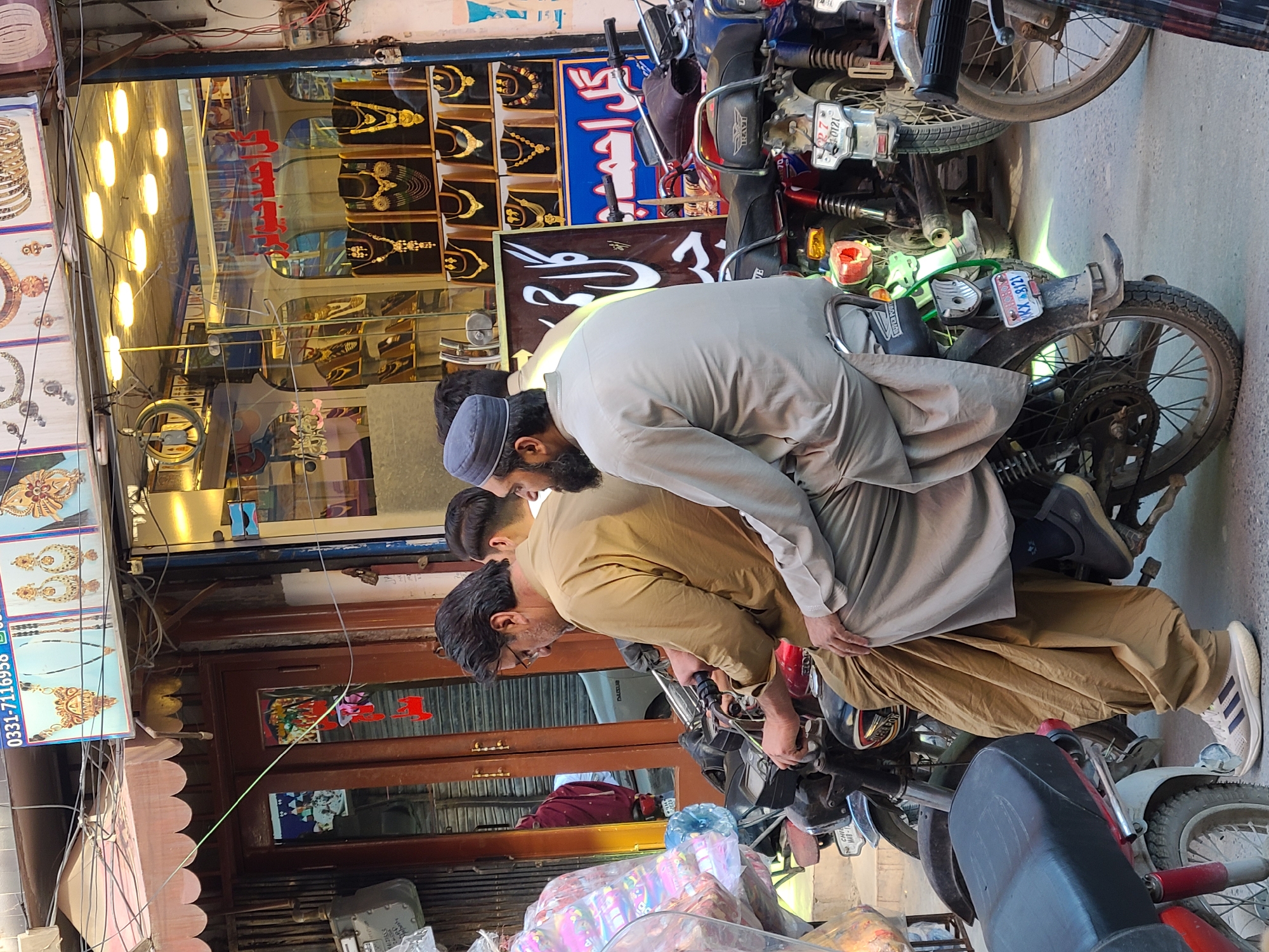 Three men are crowded around a motorcycle parked on a city street in Quetta City Tehsil, Pakistan. The motorcycle has a license plate that reads "KY-8121". The man closest to the motorcycle wears a tan shirt and pants, and white Adidas sneakers. A second man, wearing a grey outfit and a grey cap, is slightly behind him. A third man is between them. In the background, a shop selling gold jewelry is visible, with bright lights illuminating the display. Another motorcycle is parked nearby, obscuring the wall on that side. The atmosphere suggests a bustling commercial area.