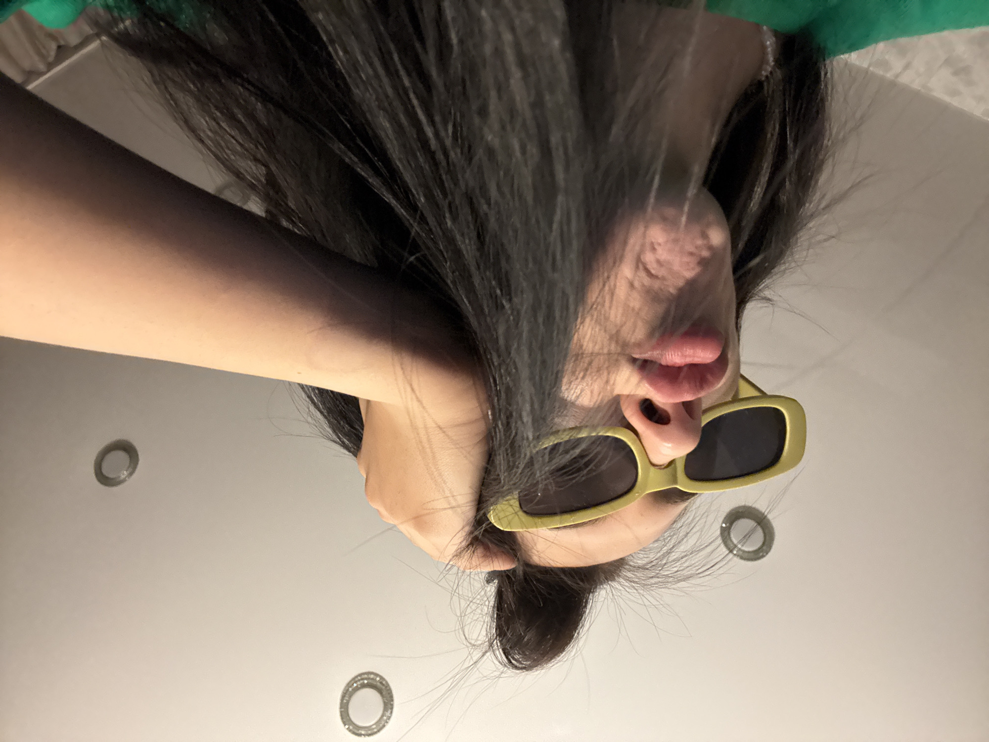 A woman with long dark hair and wearing sunglasses is lying on her back on a white surface, possibly a bed or floor. Her left arm is bent and resting on the surface, near her head. The sunglasses have a yellow frame and dark lenses. Three recessed lighting fixtures are visible in the background. The location is not discernible from the image.