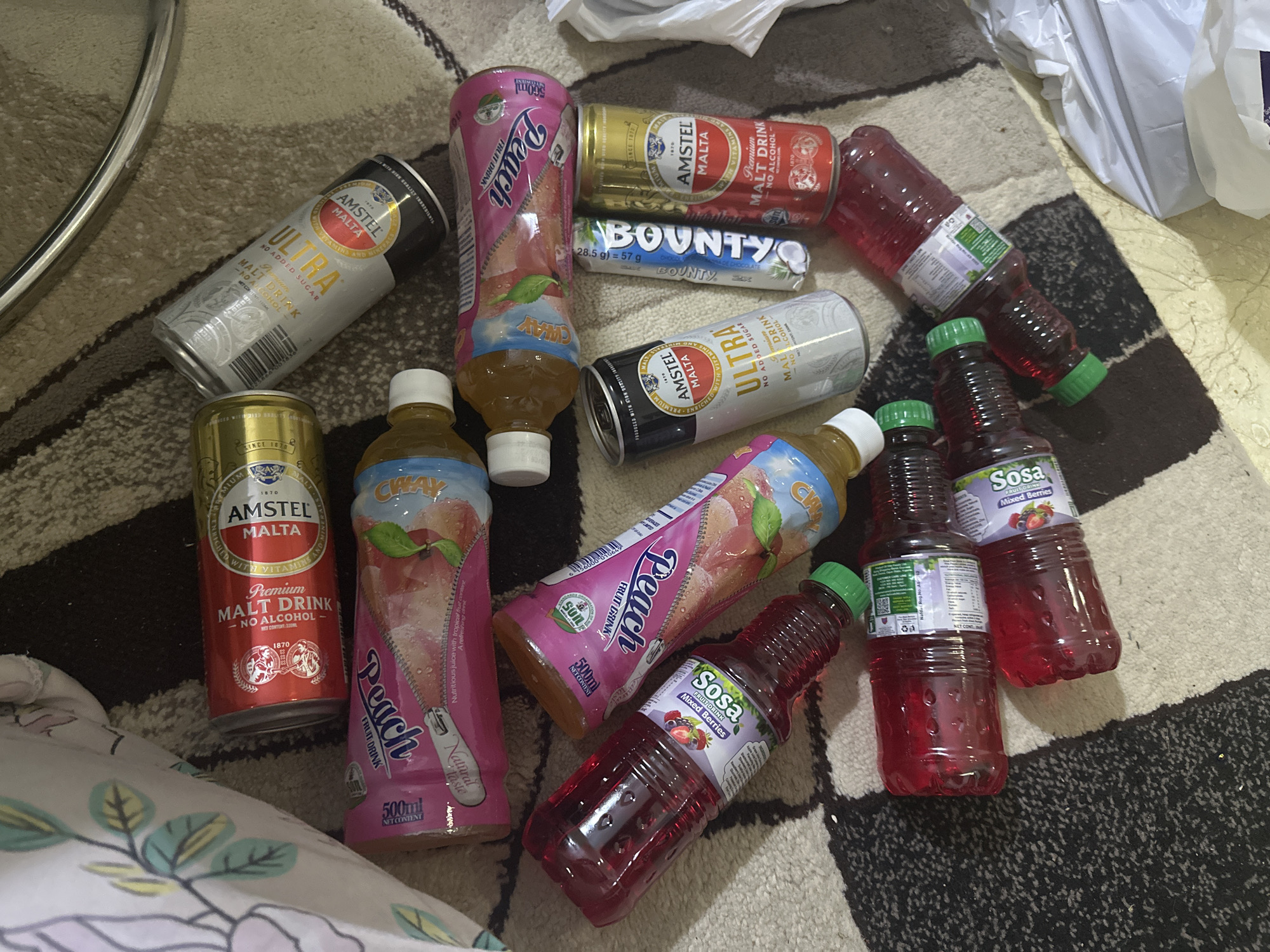 The image displays a collection of beverages, including cans of Amstel Malta (red and gold, and silver "Ultra" varieties), bottles of Peach fruit drink, and bottles of Sosa mixed berries drink. A Bounty chocolate bar is also visible. The items are scattered on a patterned carpet, possibly in a residential setting. No people or actions are depicted, and the location in Rido, Nigeria, is not visually discernible from the image itself. The lighting suggests an indoor environment.