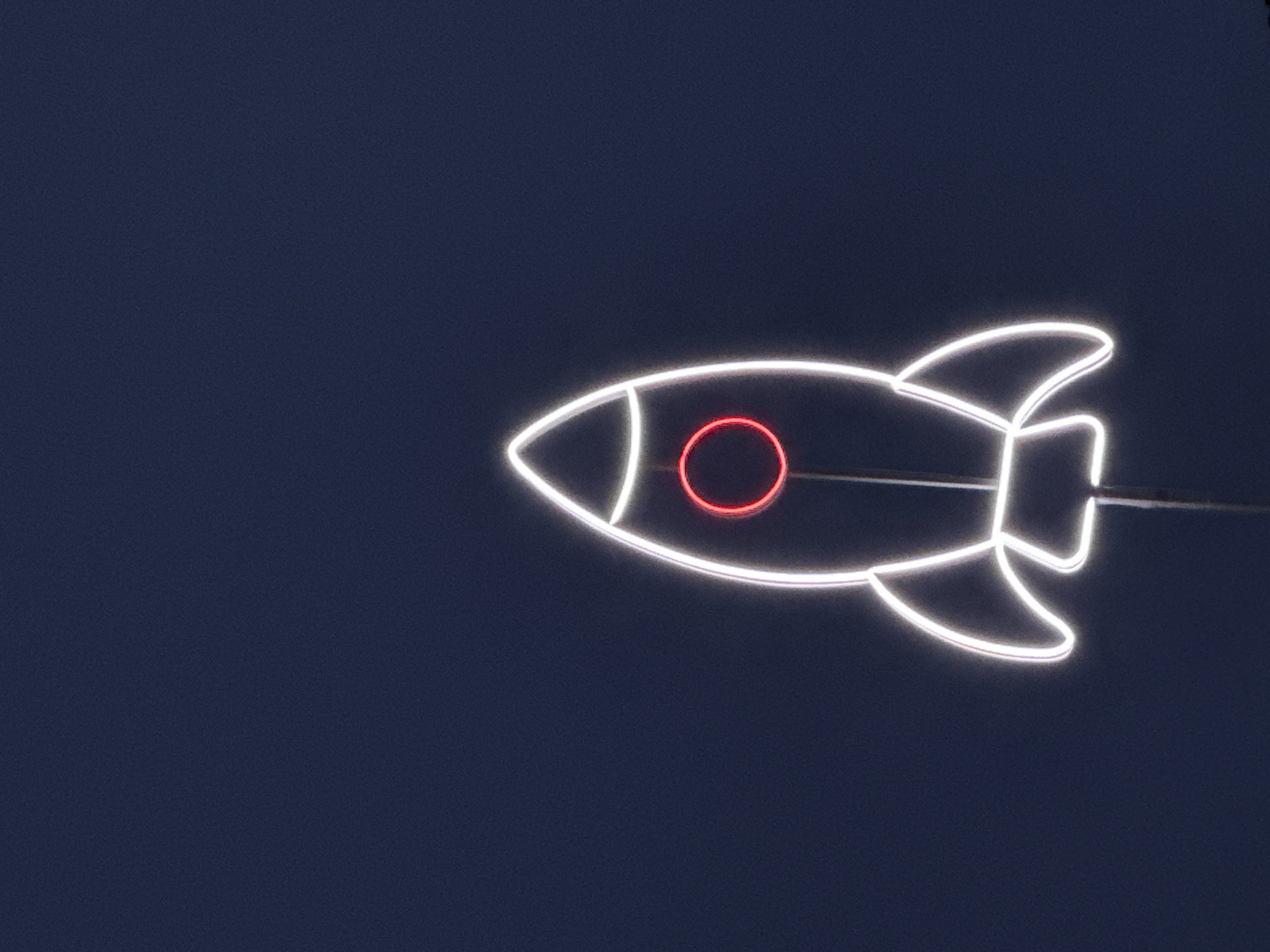 A nighttime shot shows a neon sign shaped like a rocket, outlined in white, with a red circle in the middle, set against a dark blue, almost black, background. The rocket is oriented horizontally. A thin wire cuts across the top of the rocket, slightly obscuring the upper part of the tail fin. The image appears to be taken at night due to the darkness and the brightness of the neon. It's not possible to confirm the location being Chaikovsky, Russia from the visual data.