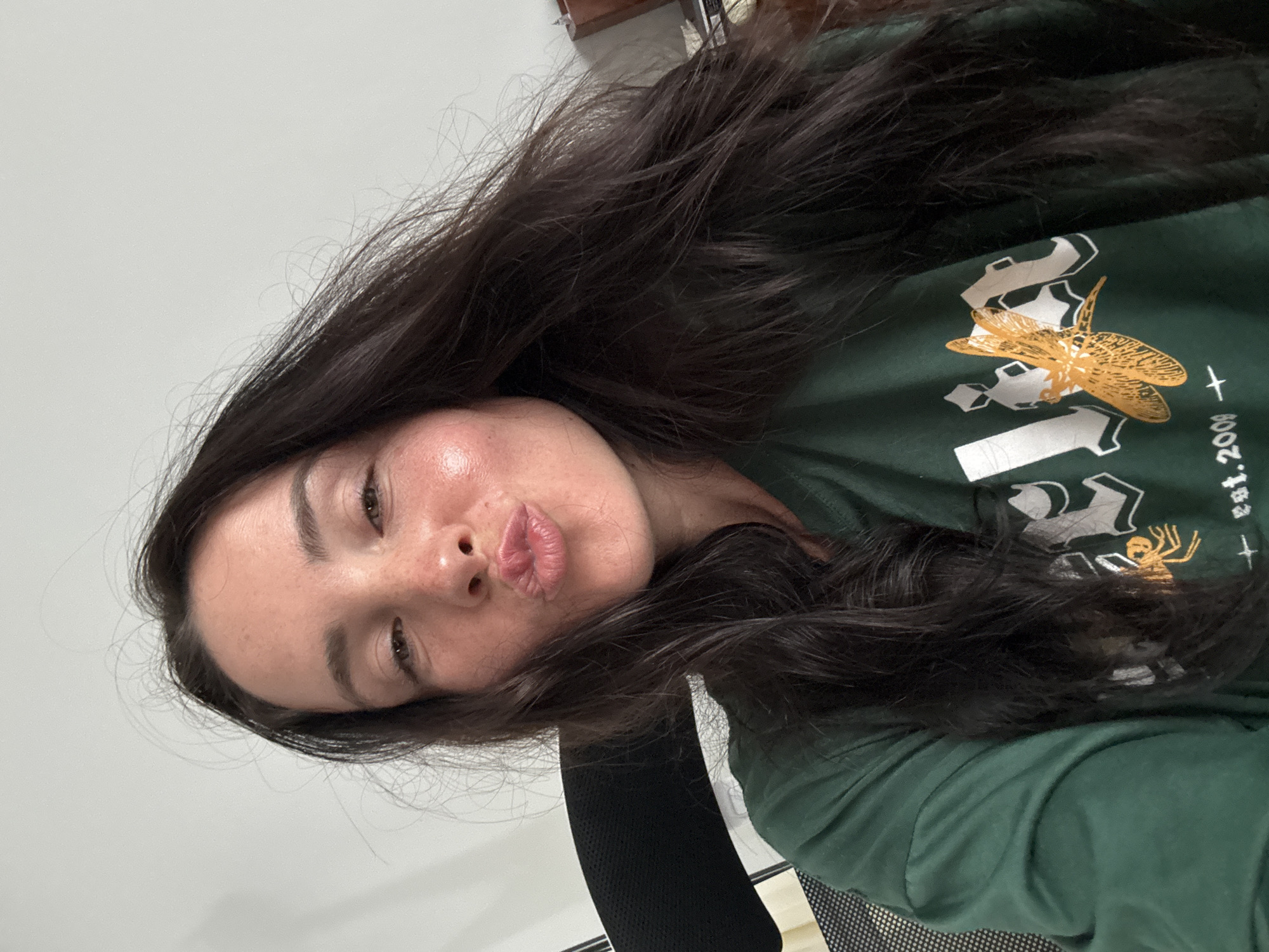 A selfie is taken by a woman with long, dark hair and light skin, showing her face from the nose up. She is wearing a green t-shirt with a graphic design on it. The design features a yellow dragonfly and what appears to be a spider. There is also text on the shirt that is partially visible. The woman is making a kissing face, and her cheeks are flushed. Her eyes are open and looking towards the camera. She has freckles on her nose and cheeks. The background is a plain white wall. The lighting suggests it is daytime, and the setting appears to be indoors, possibly an office or home environment. There are no obvious cues to indicate the weather or a specific time of day beyond it being daylight. The woman's expression conveys playfulness or affection. The visible text on the shirt reads "est. 2008".