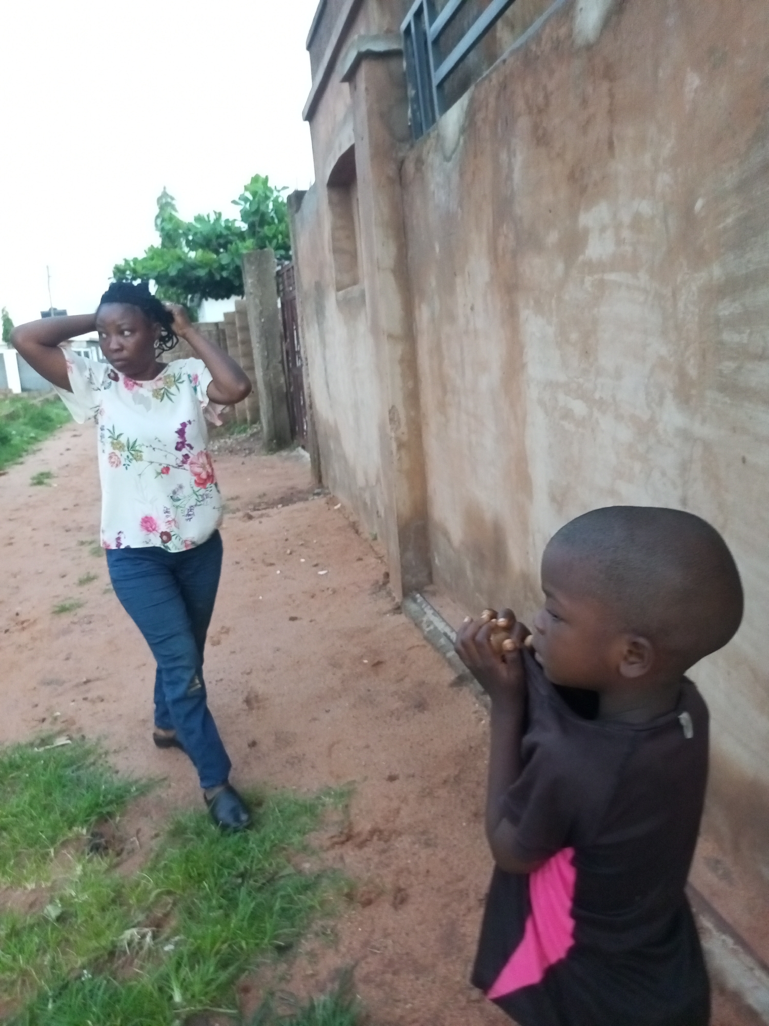 In Tempure, Nigeria, a young child with a shaved head stands near a textured, tan-colored wall, facing right with hands clasped near their chin. The child wears a dark t-shirt with a pink accent. To the left, a woman with dark hair pulled back is adjusting her hair with both hands. She wears a white t-shirt with a floral pattern and dark blue jeans. The ground is a dirt path with some patches of green grass. A building with a metal railing is visible in the background.
