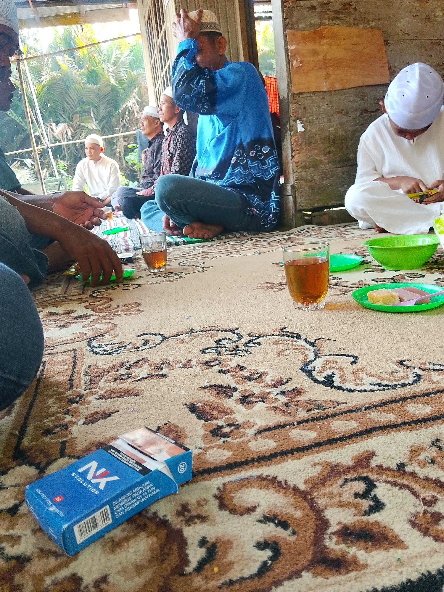 The image depicts an indoor gathering, likely in a residential setting in Buluan, Indonesia, given the context. Several individuals, predominantly male and wearing traditional attire including peci (caps), are seated on a patterned carpet. The atmosphere appears relaxed, with some individuals engaging in conversation or observing their surroundings.
In the foreground, a pack of "NK Evolution" cigarettes is prominently displayed on the carpet. Nearby, a glass of amber-colored liquid, likely tea, sits next to small green plates holding what appear to be sweets or snacks. A green bowl and another small green plate are also visible. The lighting suggests it is daytime, with natural light filtering in from an open doorway or window in the background, revealing lush greenery outside. The walls of the structure are made of rough-hewn wood, and a portion of a tin roof is visible. The overall scene conveys a sense of casual social interaction and hospitality.