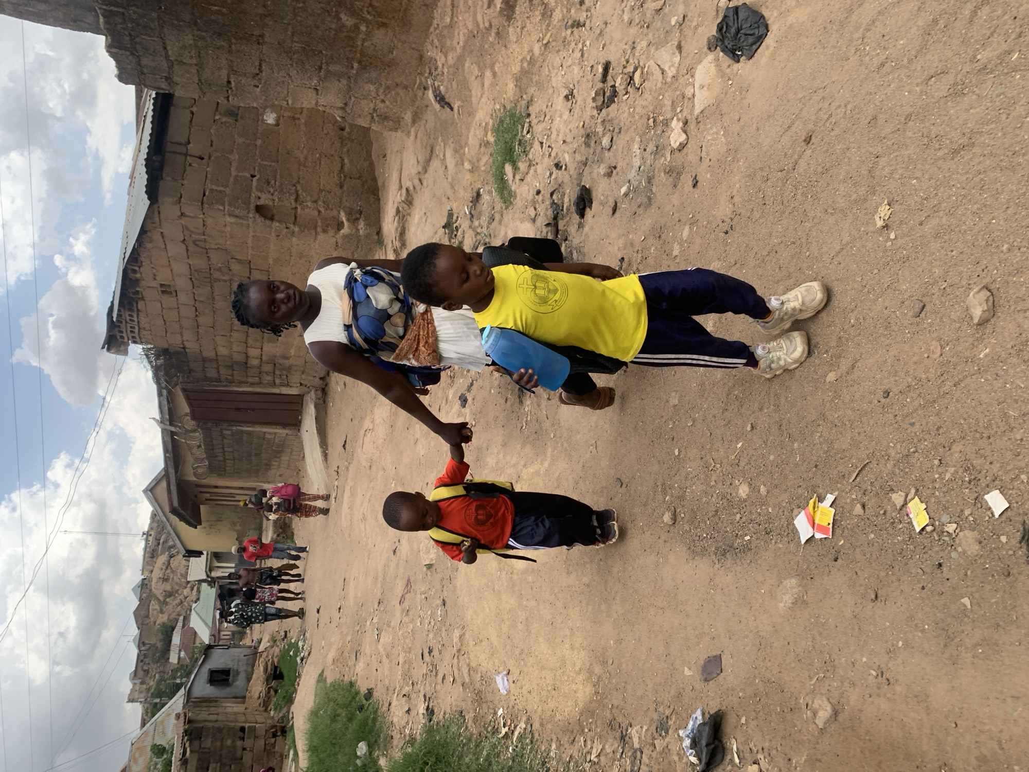 In the city of Jos, Nigeria, a woman is walking with two young children on a dirt road. The woman is holding the hand of the child on the right, who is wearing a red shirt, dark blue pants, a yellow backpack, and black shoes. The second child on the left is wearing a yellow shirt, dark blue pants with a white stripe, a black backpack, and white shoes. The area around them consists of buildings constructed from bricks and corrugated metal roofs. In the background, more people are walking in the same direction. The environment is dusty, with some scattered litter on the ground.
