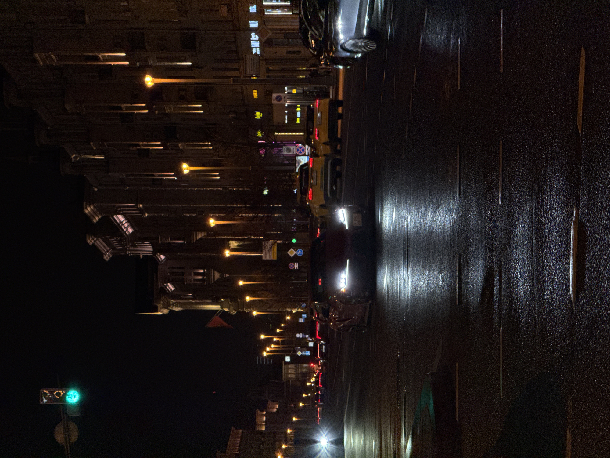 The image depicts a nighttime street scene in Moscow, Russia. The view is of a wet asphalt road reflecting the lights of passing vehicles and street lamps. Buildings line the left side of the road, illuminated by ornate streetlights. A traffic light displays a green signal. Cars and taxis are visible on the road. Street signs are present along the buildings.
