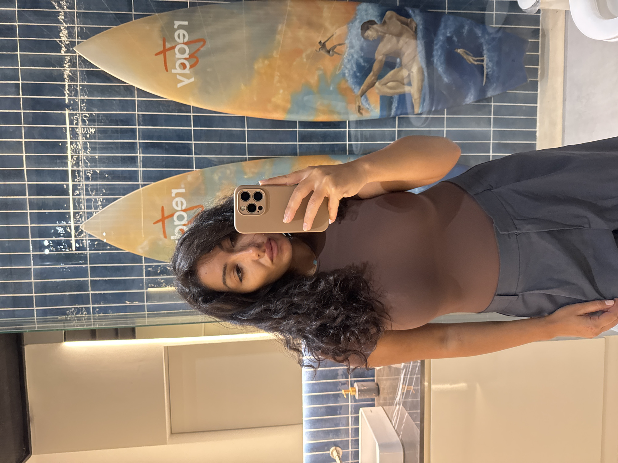 In a mirror selfie taken indoors, a woman with dark, curly hair and a neutral expression is captured. She holds a gold-colored smartphone (an iPhone with three visible cameras) up to a mirror, through which the photo is taken. She is wearing a form-fitting brown top and grey pants. A delicate light blue necklace is visible around her neck.
The background, reflected in the mirror, reveals a wall covered in dark blue rectangular tiles, creating a grid pattern. Two decorative surfboards are mounted on this tiled wall. The upper surfboard displays an artistic depiction of a male figure in a classical style, possibly nude, interacting with waves, with other smaller figures or elements in the background. The lower surfboard, partially obscured by the woman, shows a similar, though less clear, design.
On both surfboards, a brand name or logo is visible, appearing reversed due to the mirror reflection. The visible text reads "qba9l", which would be "Leadq" when un-reversed. Below the main mirror, a light-colored vanity or counter area with a sink and a grey soap dispenser with a gold pump is visible, further indicating a bathroom or changing room setting. The overall aesthetic suggests a modern, possibly resort-style, interior. The location context provided is Kuta Selatan, Indonesia, which aligns with the surf-themed decor.