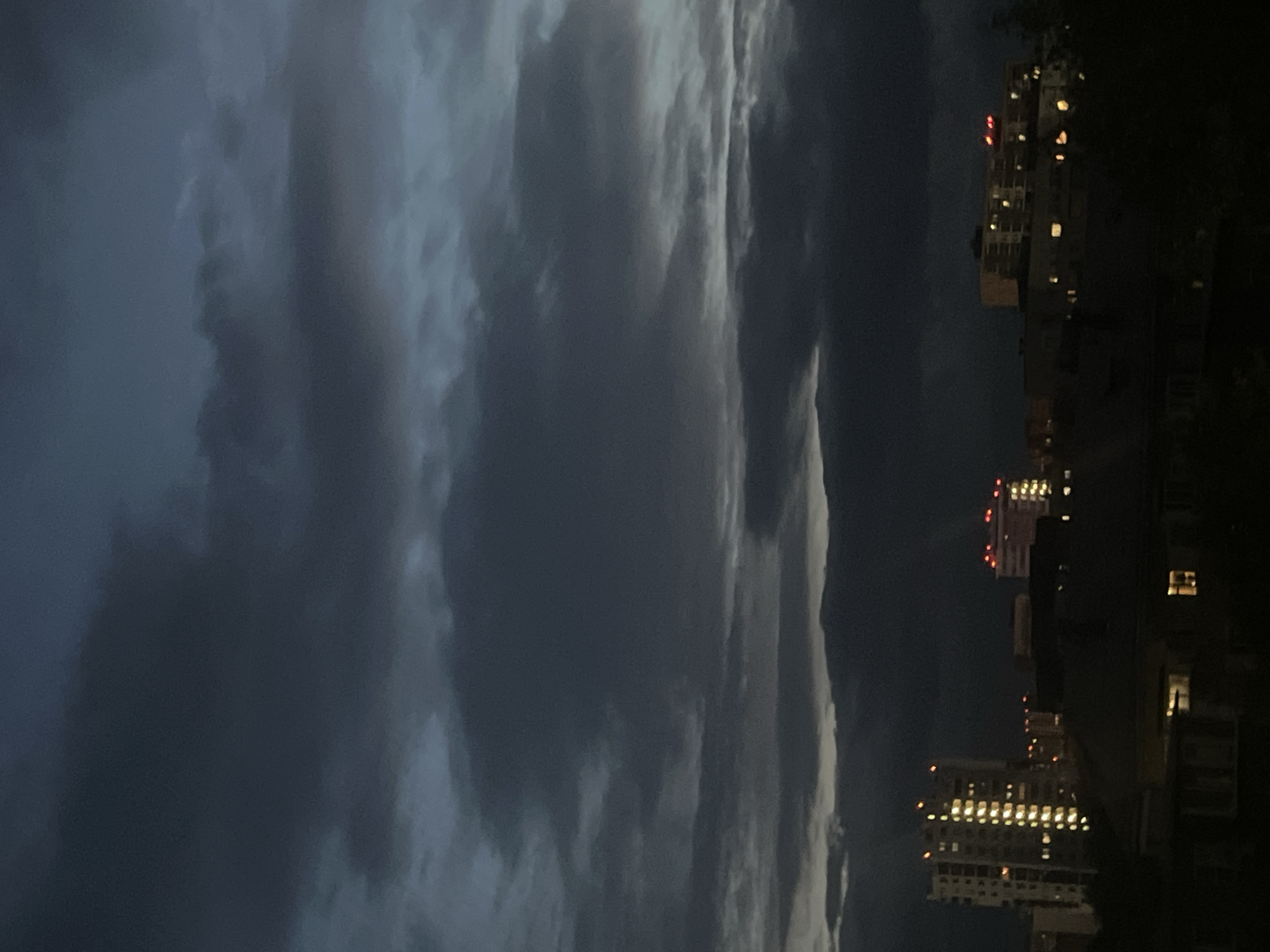 The media file displays a nighttime scene with a dramatic sky over a city. The sky is filled with dark, textured clouds, suggesting either impending weather or the aftermath of a storm. The lighting is dim, indicative of either dusk, dawn, or a heavily overcast night.
On the right side of the frame, several buildings are visible, lit from within by warm yellow lights from their windows. These appear to be apartment buildings or hotels, characteristic of an urban environment. Some buildings have red lights on their upper levels, possibly indicating navigation lights or warning signals. The overall impression is one of a city at night, with the natural drama of the sky contrasting with the man-made structures. There is no visible text in the media file. The location context provided is Chelyabinsk, Russia, which fits with the urban setting.