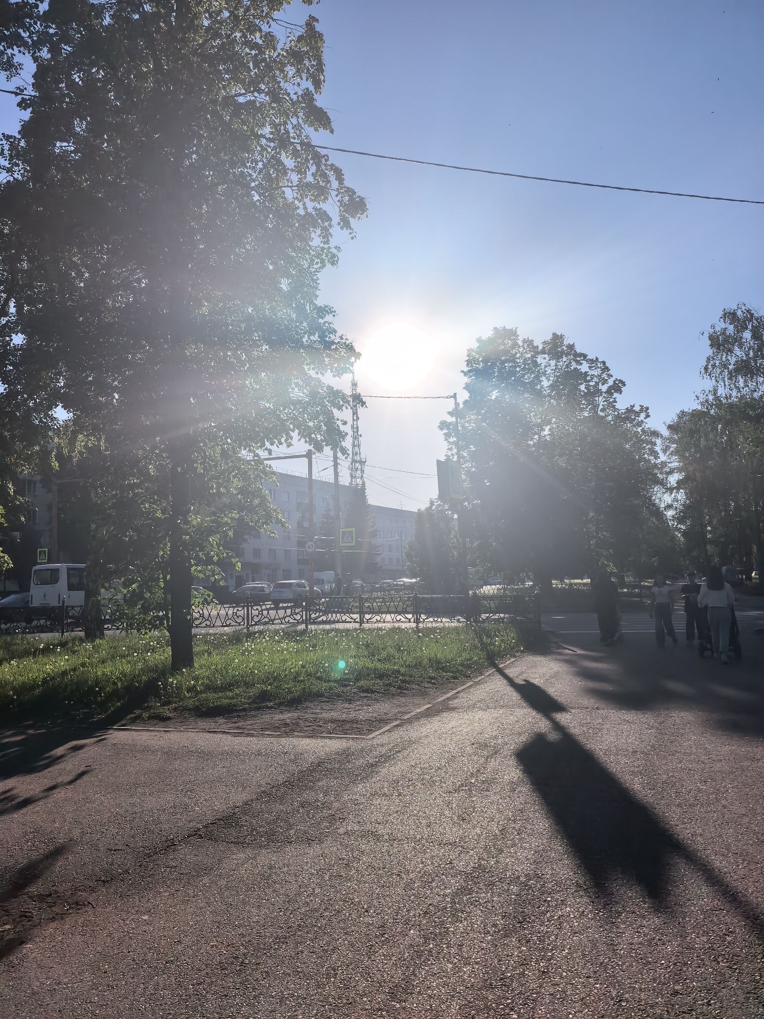 The image depicts a bright, sunny day in an urban area, likely Sterlitamak, Russia. A paved road stretches into the distance, marked with shadows of trees and pedestrians. A group of people, possibly a family, is walking on the right side of the road. On the left, there is a patch of grass with dandelions, a fence, and cars. Buildings are visible in the background, partially obscured by trees. The sun is shining brightly in the sky, causing lens flare. There is a utility pole with a cable crossing the sky. There's a pedestrian crossing sign visible behind the trees and the fence.