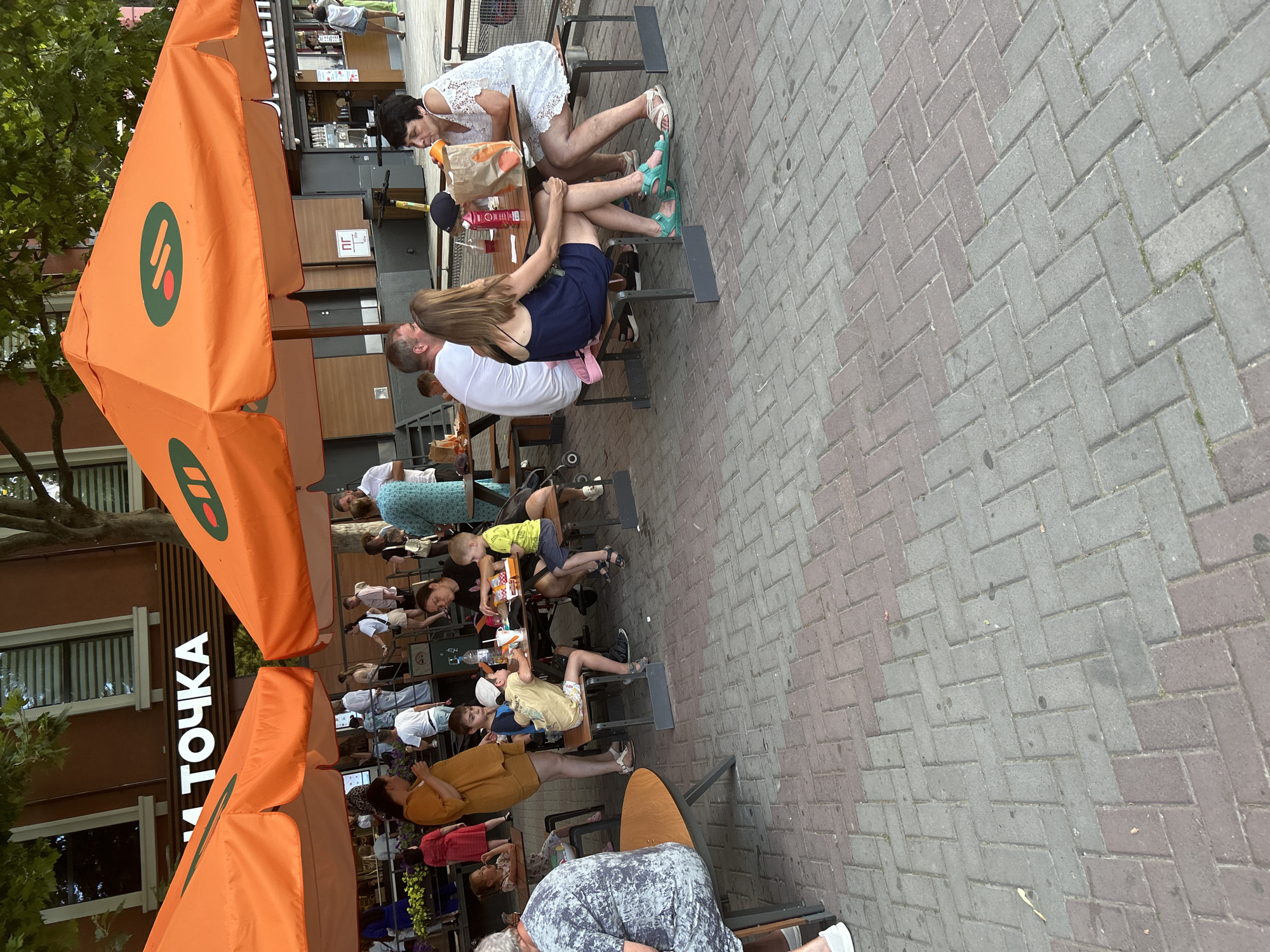 A group of people are dining at outdoor tables under orange umbrellas. The tables are made of wood with metal legs. The ground is paved with brick-like stones. In the background, a building has the word "ТОЧКА" in large white letters above a window. The scene appears to be a casual dining area in a city. This specific location is likely a street or square in Gelendzhik, Russia, as indicated by the text on the building, which reads "ТОЧКА" (TOCHKA), a common sign for a cafe or fast food establishment in Russia, and the overall aesthetic is consistent with establishments found in Russian resort towns like Gelendzhik.
The people visible include adults and children. One group consists of a woman in a white dress and a man in a white shirt, sitting at a table. To their right, a woman in a blue dress is seated, and another woman in a white dress is visible further to the right. In the foreground, a woman in a mustard-yellow dress stands, and a young boy in a yellow t-shirt and a cap is seated at a table. Another boy in a blue shirt is also at the table. A person in a grey and white patterned shirt is seated at a table to the left. Other individuals are visible in the background, some walking and some standing. The umbrellas have a green and red logo. The environment is outdoors, with some trees providing shade.