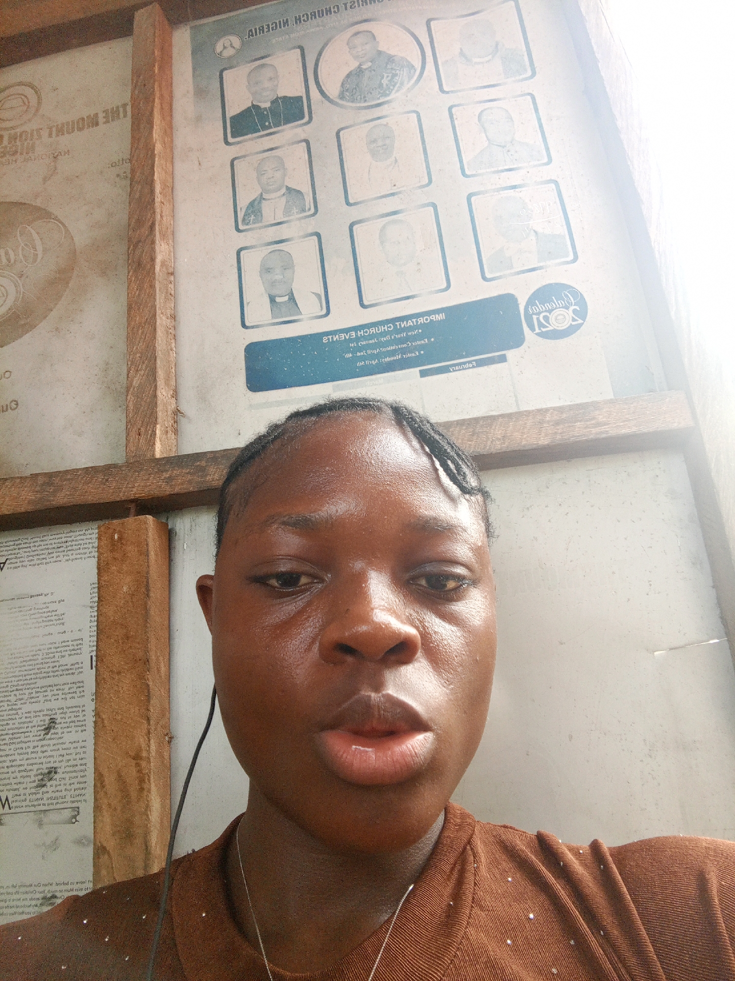 The image displays a close-up selfie of a young woman in an indoor setting. She has dark skin, her hair is styled in small braids, and she looks directly at the camera with a neutral expression. She is wearing a brown, textured top adorned with small, sparkly embellishments, and a thin silver necklace. An earbud cord is visible near her left ear and runs down her front.
Behind her, the background consists of wooden planks forming part of a structure, and two posters or signs attached to a wall. The more prominent poster on the right features a grid of nine black-and-white portraits of men, likely clergy, dressed in ecclesiastical attire. The text on this poster is mirrored, but several phrases are discernible, including "CHRIST CHURCH NIGERIA," "IMPORTANT CHURCH EVENTS," and the year "2021." Other partially visible mirrored text includes "Feburary" and references to dates (e.g., "Jan," "April"), suggesting a calendar of events. On the left side, another mirrored poster displays the words "THE MOUNT ZION."
The scene is brightly lit, indicating it is daytime. The presence of the church-related posters strongly suggests the setting is within a church or religious institution, consistent with the provided location context of Mbribit, Nigeria. The image captures a personal moment against a backdrop of religious information.