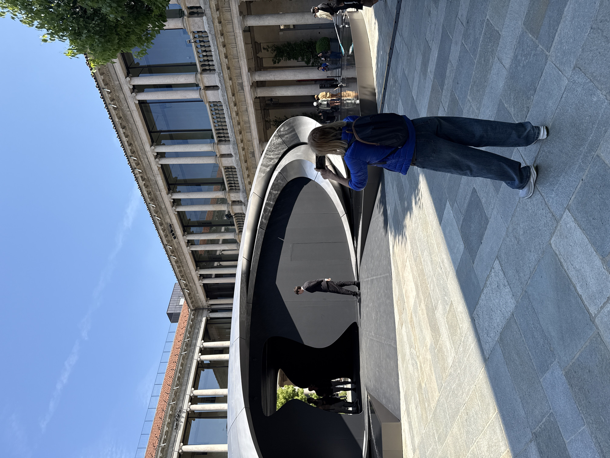 The image captures a bright, sunny outdoor scene in an urban public square, consistent with a location in Milan, Italy. In the foreground, a prominent, dark, sculptural installation with curved, overlapping metal forms takes center stage. A person dressed in black is visible standing within the shadowy, introspective interior of this modern art piece.
To the right, a woman, viewed from behind, is actively engaged with her smartphone, seemingly capturing a photograph or video of the sculpture. She is wearing a blue puffer jacket, blue jeans, and white sneakers, with a dark backpack resting on her shoulders.
The background is dominated by a grand, classical-style building featuring a long colonnade of light-colored pillars, large rectangular windows that reflect the clear blue sky, and intricate architectural details along its facade. A lush green tree partially frames the top-left corner of the building. Further back, several other individuals are visible near a shallow water feature or fountain situated in front of the historic building.
The ground is paved with light grey, rectangular stone tiles, exhibiting strong, distinct shadows cast by the sculpture and people, indicating bright mid-day sunlight. The sky above is a vibrant, clear blue with a few faint, wispy clouds. No discernible text is visible anywhere in the image. The scene portrays a casual atmosphere of urban exploration and appreciation of art and architecture.