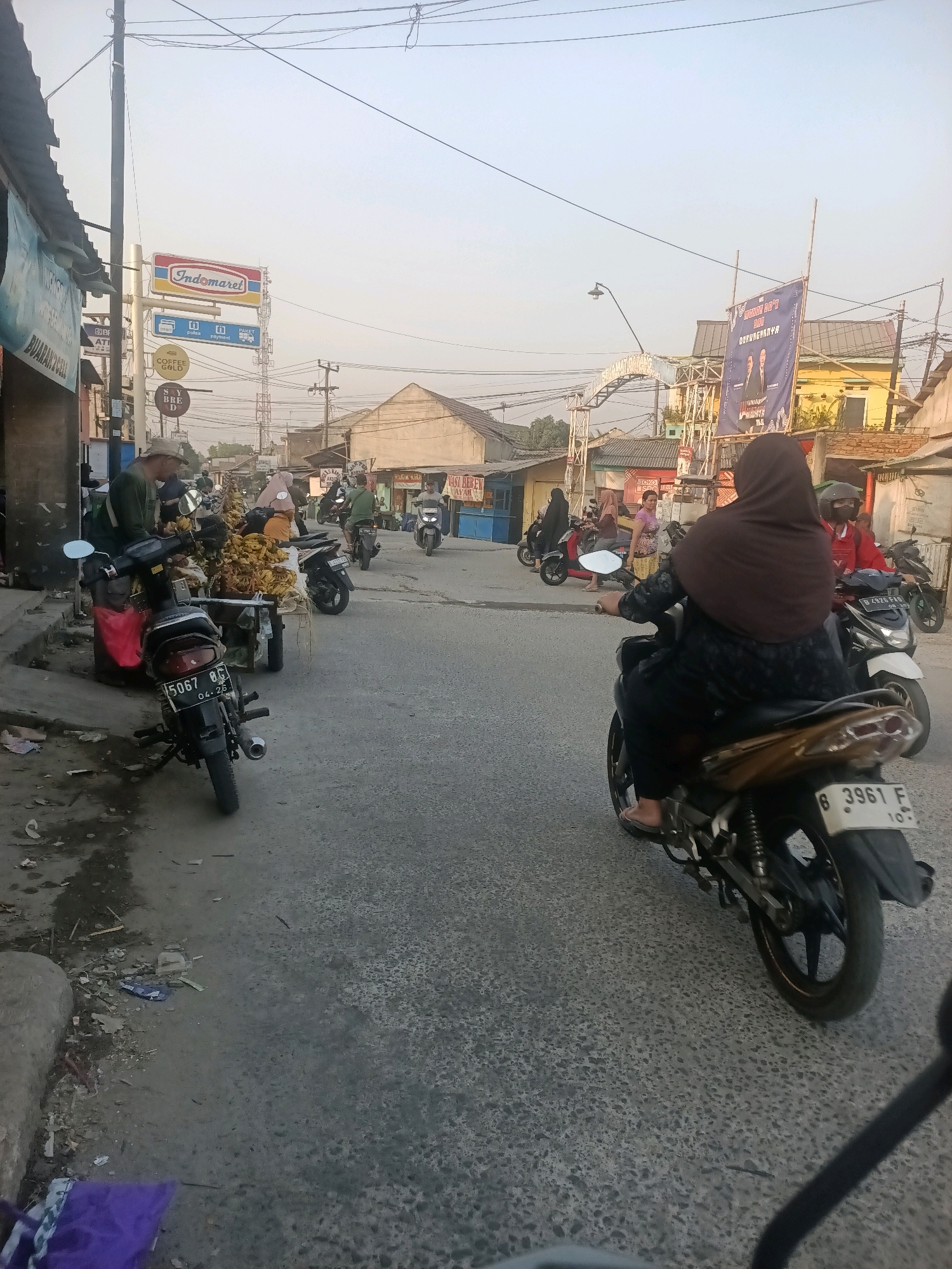 In Wanajaya, Indonesia, a street scene depicts a moderate level of activity on a paved road. Several motorcycles are present, with some in motion and others parked. In the foreground, a person is riding a motorcycle, wearing a brown hijab and a dark patterned outfit. Another motorcycle is parked to the left, displaying license plate number 5067 BC. Further down the road, more motorcycles and people are visible, suggesting a bustling local area. Buildings line the street, including one with an "Indomaret" sign, indicating a commercial presence. A large banner with political figures is also displayed. The environment appears to be an urban or semi-urban setting with overhead wires and street lighting. The ground is asphalt, with some debris and puddles present. The lighting suggests it is daytime, likely late afternoon given the soft light.
