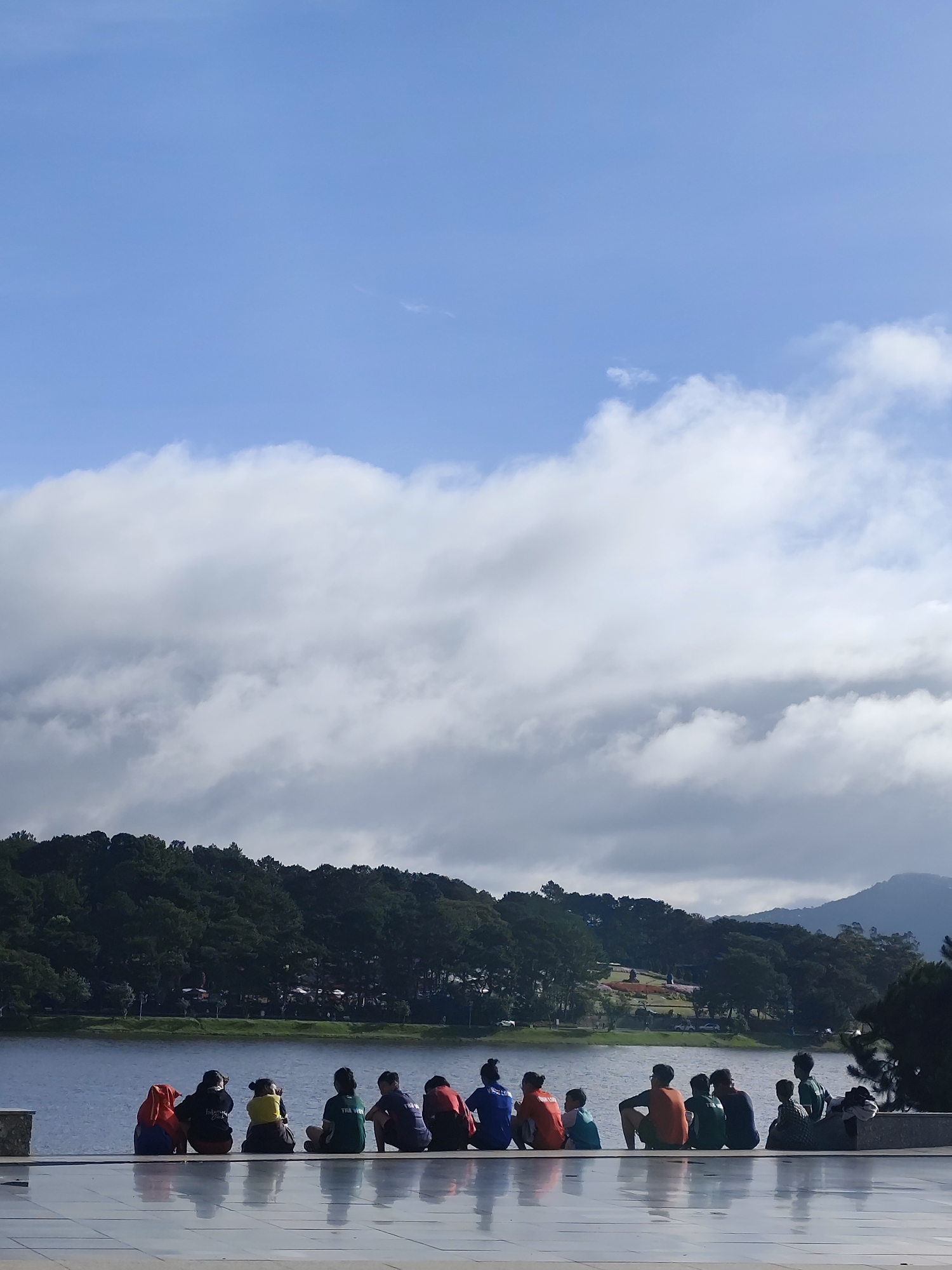 In Da Lat, Vietnam, a group of people sits on a raised concrete platform facing a serene lake. The platform has a reflective, glossy surface, creating mirror images of the individuals seated there. The individuals are dressed in various colored casual clothing, including a red hooded jacket, a black hoodie, a yellow shirt, a green t-shirt, and a blue t-shirt. They appear to be engaged in conversation or simply enjoying the view.
Behind them, the lake stretches out, its surface rippling gently. Across the lake, a dense line of dark green pine trees forms a natural border, with hints of buildings and structures interspersed within the foliage. Beyond the treeline, rolling hills and mountains, some of them misty, rise in the background. The sky above is a vibrant blue, punctuated by large, puffy white clouds that cast shadows on the landscape below. The overall atmosphere is peaceful and tranquil, suggesting a popular scenic spot in Da Lat, likely Xuan Huong Lake, known for its natural beauty and recreational activities.