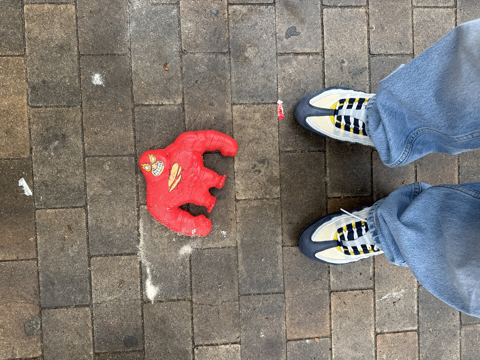 A person stands on a paved walkway made of rectangular gray pavers. The person is wearing light blue jeans and gray, white, and yellow sneakers. To the left of the person, on the pavement, is a damaged red rubber toy in the shape of a figure, with visible painted details of a face and other markings. A small piece of red and white trash lies next to one of the person's shoes. The pavers appear somewhat weathered. The environment is not identifiable as Barcelona, Spain.