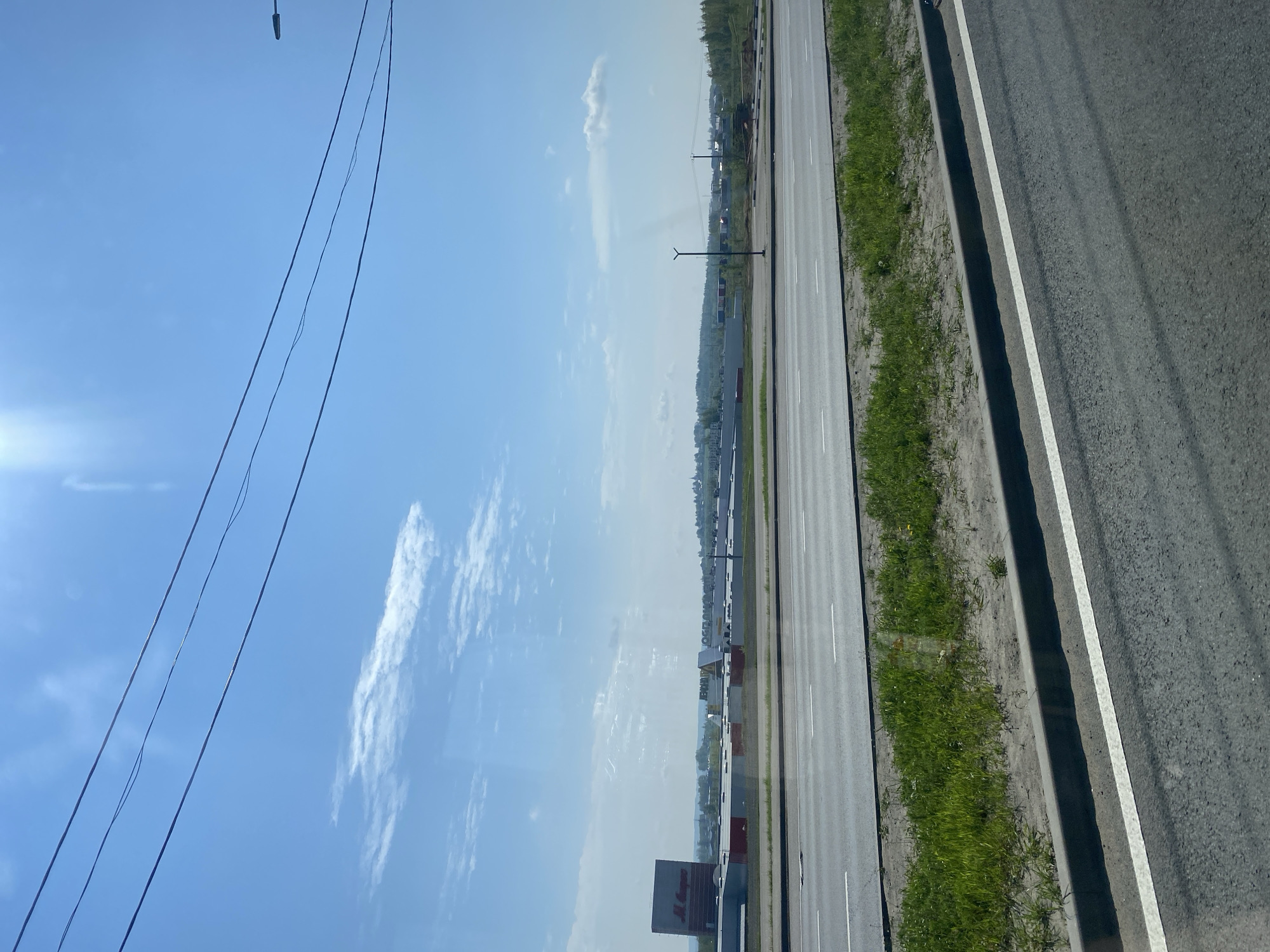 This image, set in Rusanovo, Russia, captures an outdoor scene from what appears to be a moving vehicle, looking across a multi-lane road towards a developed area. The perspective is horizontally oriented, showing a wide expanse of sky on the left and road infrastructure on the right.
The scene features a wide asphalt road with clear white lane markings, flanked by a grassy verge and a concrete curb. Beyond this, another, possibly narrower, road or parking area is visible. In the upper left portion of the bright blue sky, several dark utility lines or power cables stretch across the frame, partially obscuring a bright lens flare from the sun. The sky is mostly clear with scattered white cumulus clouds.
Along the horizon on the right side of the image, numerous low-rise buildings, possibly commercial establishments or warehouses, line the road. A modern black street light stands prominently near the road. In the foreground, near the bottom right, a large billboard-like sign with red lettering is partially visible, displaying the text "АЛМАЗ". Further in the distance, more buildings and patches of green vegetation can be seen, hinting at a suburban or exurban landscape.
No specific activity or people are visible; the image provides a static snapshot of the environment. The bright sun and clear sky indicate it is daytime, likely a clear and sunny day.