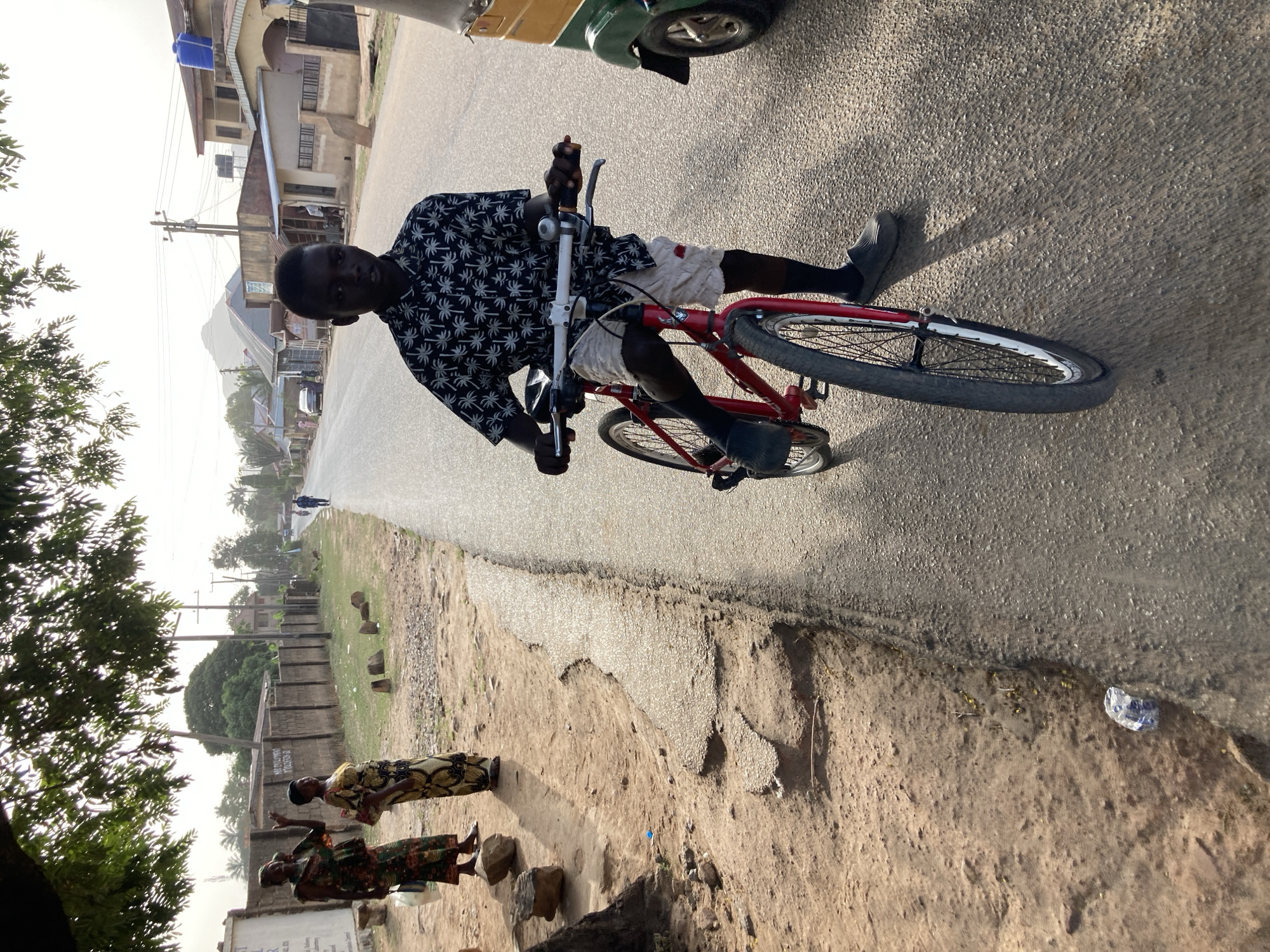 A man rides a red bicycle on an asphalt road in Jos, Nigeria. He wears a patterned shirt, light-colored shorts, black socks, and black slip-on shoes. His skin is dark, and his hair is short and dark. In the foreground, two women in traditional clothing stand along the roadside on the dirt shoulder. The background includes residential buildings and utility poles along the road, with a mountain visible in the distance. The lighting suggests daytime with diffused sunlight.