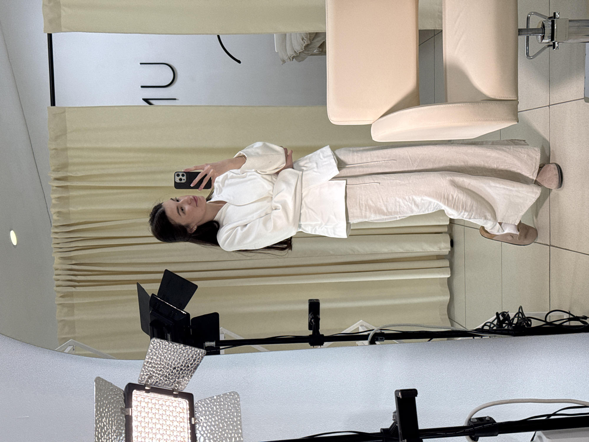 An indoor, mirror-reflection image depicts a woman taking a selfie with a smartphone, likely in a salon or studio setting in Chaikovsky, Russia. The woman, with long dark hair, is positioned centrally, looking directly at her phone. She is wearing a light-colored, long-sleeved jacket or robe over matching wide-leg trousers, and light-colored slippers.
The background shows a light-colored curtain extending across the width of the frame behind her. To the right, two light-colored, upholstered chairs, possibly salon chairs, are visible. In the upper part of the reflection, above the curtain, there is a light-colored wall or partition with dark, inverted characters that, when viewed correctly, appear to be part of a word or logo, possibly "NU" or "ЛU".
In the foreground, on the side from which the photo is taken, two professional LED panel lights are visible, one illuminated, casting bright artificial light. These lights are mounted on stands, and various cables and wires are visible on the floor around the equipment. The floor is tiled in a light, neutral color. The overall impression is one of a well-lit, professional environment.