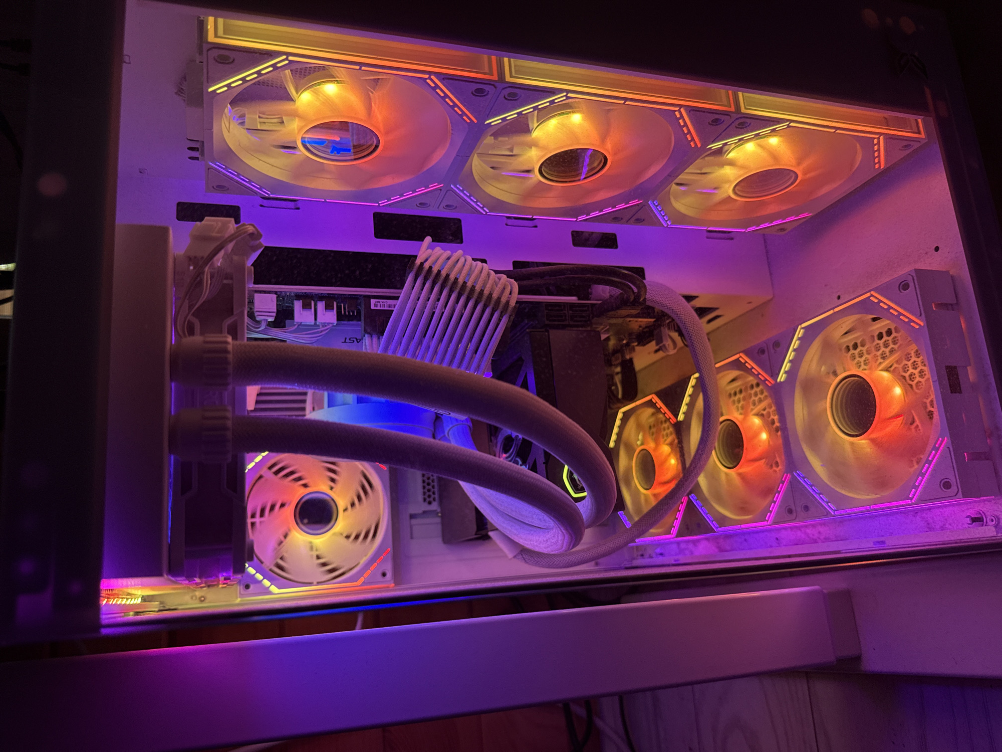 The image displays the interior of a desktop computer case, illuminated by multi-colored LED lighting. The dominant colors are shades of purple, pink, orange, and yellow.
Six fans are visible within the case. A single fan is mounted at the top-rear, exhibiting yellow and orange illumination on its blades, which show rotational blur. Three vertically stacked fans are visible on the right side of the case, likely front intakes, each displaying white blades with yellow and orange light and surrounded by LED rings featuring segments of yellow, orange, and pink. Two additional fans are mounted horizontally on the bottom of the case, showing orange illumination on their blades, which also exhibit rotational blur, and surrounded by LED rings with purple, pink, and orange segments.
A liquid cooling system's radiator is mounted at the top of the case, with two white-sleeved tubes extending downwards towards what appears to be a CPU block. A bundle of white-sleeved cables, consistent with a 24-pin ATX power cable, is visible vertically along the left side of the motherboard, secured by cable combs. Portions of a motherboard are visible behind these cables, bearing the text "AST". A graphics card, dark in color with hints of blue/purple lighting, is horizontally positioned below the radiator and above the CPU block area. The interior surface of the computer case is white, and a light accumulation of dust is present on the bottom panel.
The location of this scene in the city of Busmantsi, Bulgaria, cannot be determined from the visual information provided in the image itself. The image depicts only the internal components of a computer case.