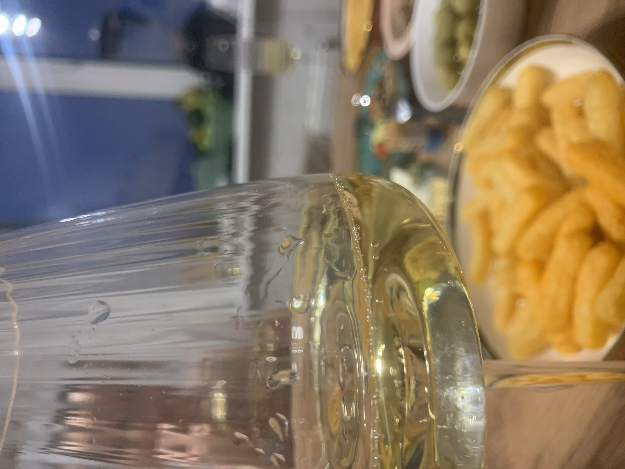 A close-up shot captures a partly-filled glass of yellow liquid with bubbles clinging to its sides. Behind the glass, there is a blurred view of what appears to be a plate filled with french fries and possibly other dishes. The background is further blurred, showing some indistinct shapes and colors. The location is not discernable.