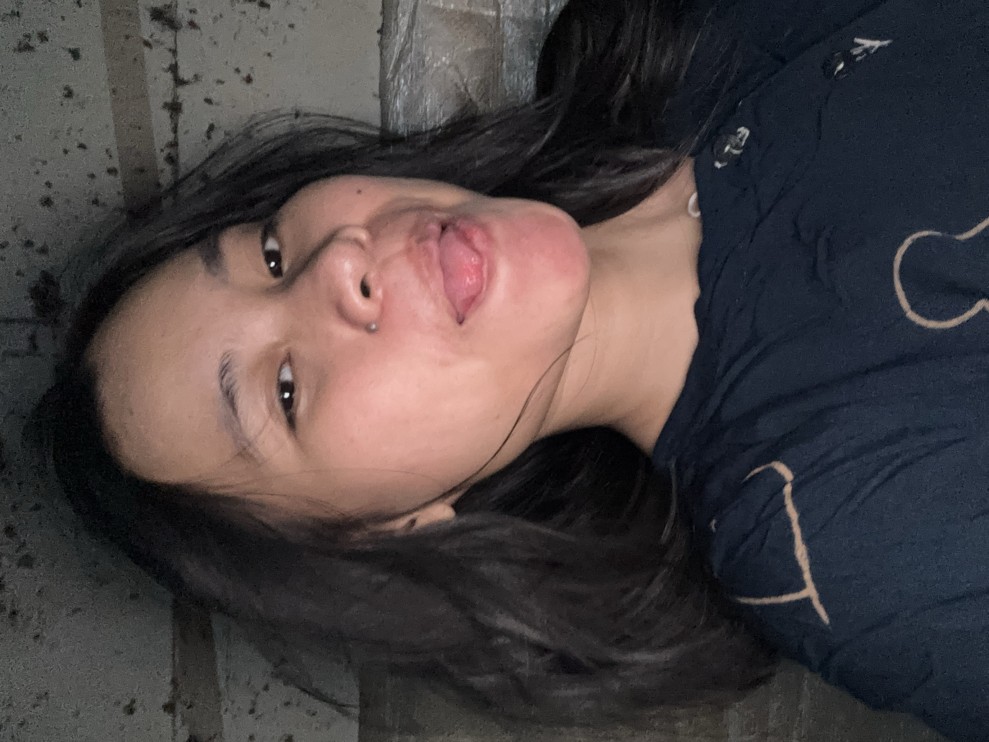 A young woman with dark hair and light brown skin is lying down, her head tilted upwards. Her mouth is slightly open with her tongue sticking out. She is wearing a dark blue t-shirt with a tan outline design on it. The background appears to be a textured, possibly concrete or tiled surface, with some darker specks scattered across it. The lighting suggests it might be indoors or nighttime, with a dim and somewhat grainy quality to the image. Her expression is playful or mischievous. The scene is oriented sideways.