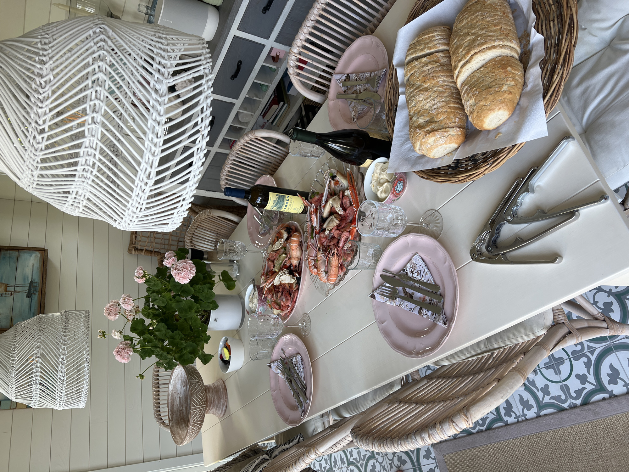 This is a high-angle, medium shot of a dining table set for a meal, likely outdoors or in a sunroom, with light filtering in. The table is made of pale wood and is laden with food and place settings. Two large platters of seafood, appearing to be shrimp and crab, are prominently displayed. Beside the seafood are small bowls of what looks like mayonnaise or aioli, and a bowl containing sliced radishes and lemon. Two bottles of white wine stand on the table, one open.
Each place setting includes a pale pink plate with a floral napkin and cutlery. There are several glasses for wine and water. In the foreground, a basket holds two loaves of sliced baguette. A nutcracker sits on the table near the bread. In the background, a potted plant with pink flowers adds a touch of color. The table is surrounded by light-colored wicker chairs. Behind the table, a wall with horizontal wooden planks is visible, along with a grey cabinet with numerous drawers. A large, white, woven pendant light fixture hangs overhead. A framed artwork depicting a lighthouse and a boat is on the wall in the upper left. The floor has a patterned tile design. The overall impression is a casual, seaside-inspired meal setting. The scene is located in Larkollen, Norway.