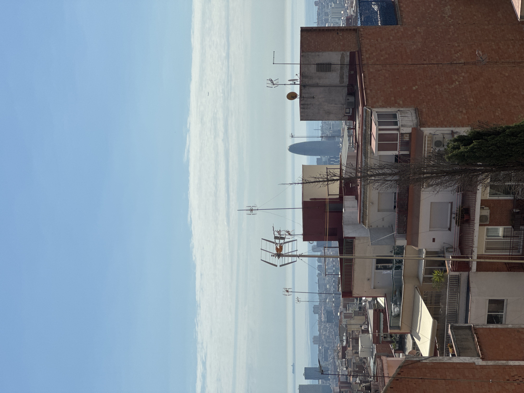 The image depicts a cityscape of Barcelona, Spain. Prominent in the distance is the Agbar Tower, a cylindrical glass structure. The foreground consists of densely packed residential buildings with red brick facades and balconies. Numerous antennas and satellite dishes are visible on the rooftops. The sky is mostly clear with a band of clouds near the horizon. The image is oriented sideways.