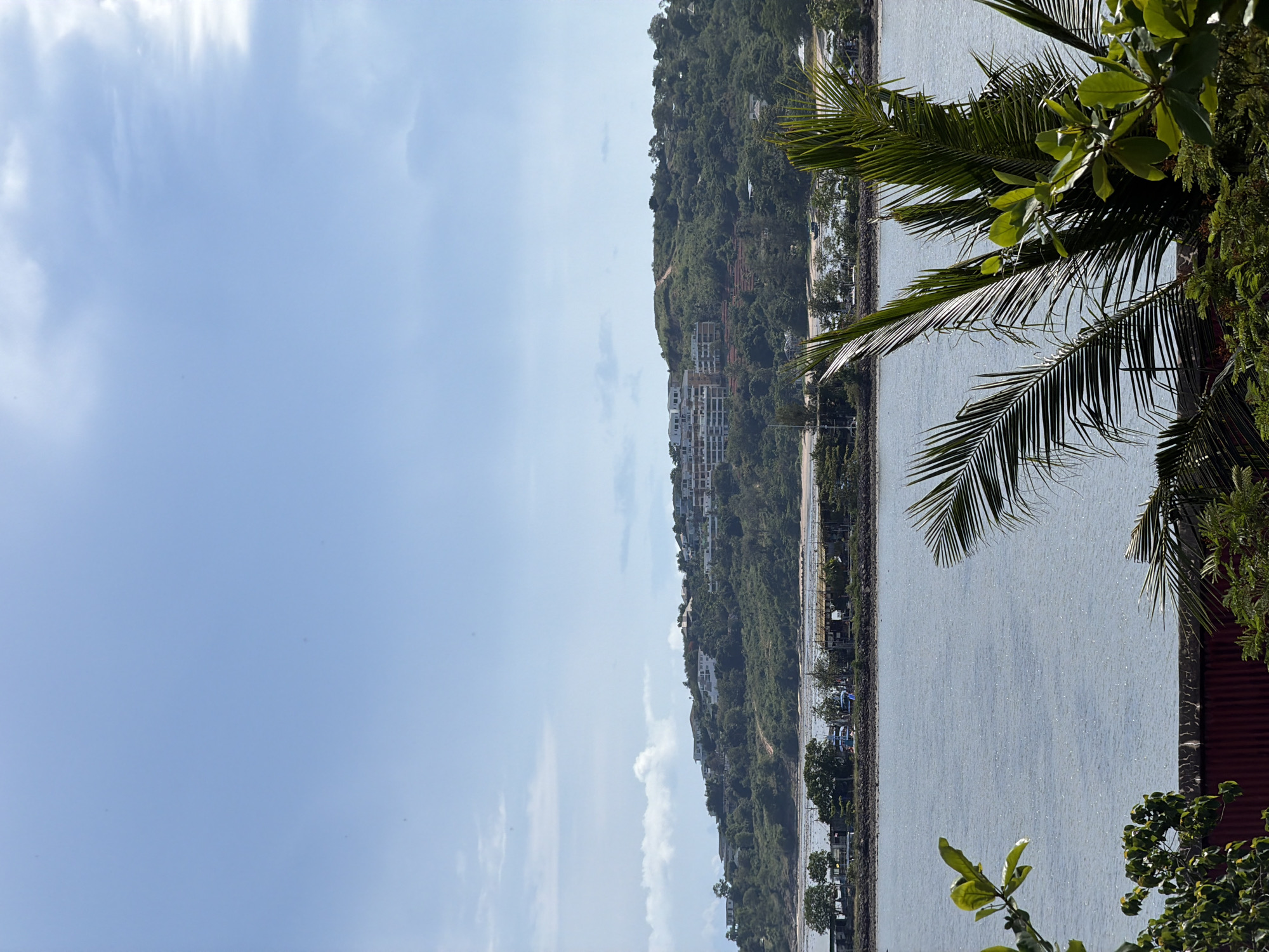 The image shows a wide shot of a coastal scene in Baan Plai Laem, Thailand. In the foreground, there are palm fronds and other green foliage, with a red corrugated metal structure visible at the bottom. A calm, blue body of water occupies the middle ground, with a rocky shoreline dotted with trees and small structures along the opposite bank. Beyond the water, a lush green hill rises, covered with dense vegetation and scattered white buildings, suggesting a residential or resort area. The sky above is a pale blue with wispy white clouds. There are no people or animals visible in the image, and no specific actions or interactions are depicted. The scene is serene and captures a typical coastal landscape with natural elements and human development.