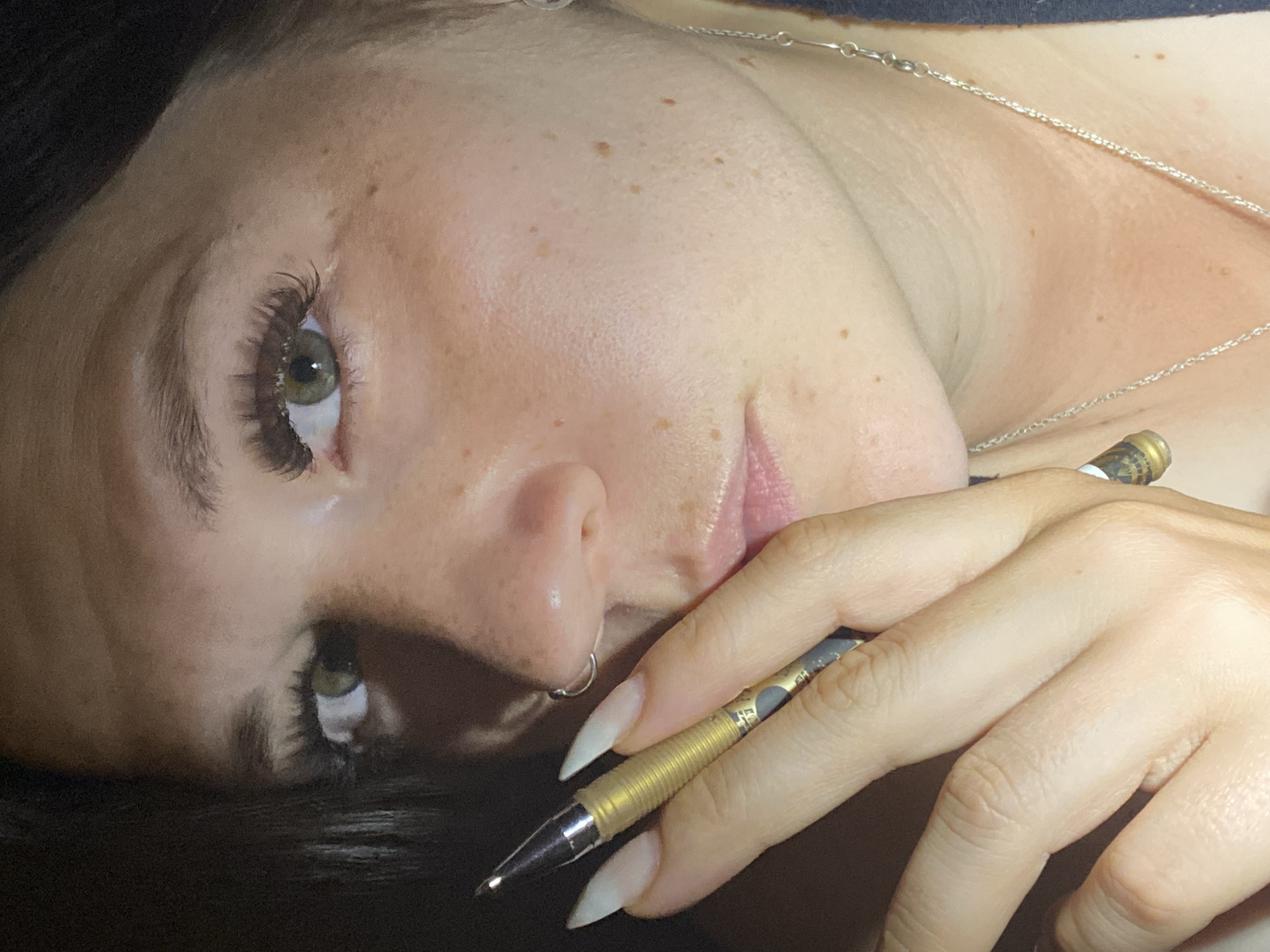 The image is a close-up, frontal portrait of a woman's face, angled slightly to the left, with her right hand holding a mechanical pencil near her mouth. She has fair skin with freckles and is wearing false eyelashes. A silver hoop earring is visible in her right nostril. A thin silver necklace is also visible around her neck. The background is dark and indistinct, suggesting an indoor setting. There is no discernible connection to Chaikovsky, Russia, in the visual information provided.