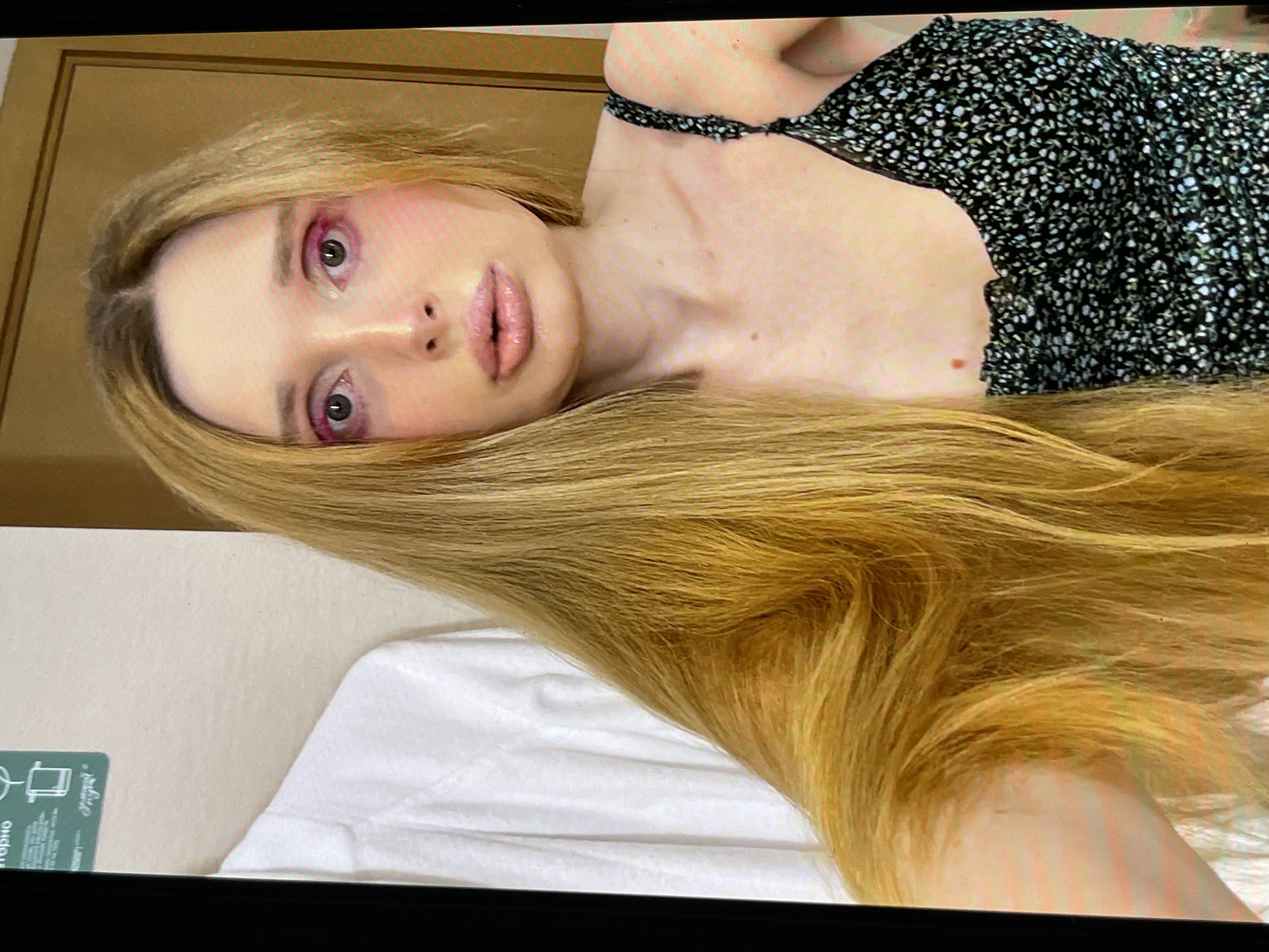 A young Caucasian woman with long blonde hair, heavy pink eye makeup, and plump lips poses for a selfie in an indoor setting. She is wearing a black floral print dress with thin straps. To her left, a white garment, possibly a shirt, hangs on a rack. The background features a wall and a wooden door. The image is taken from the chest up.