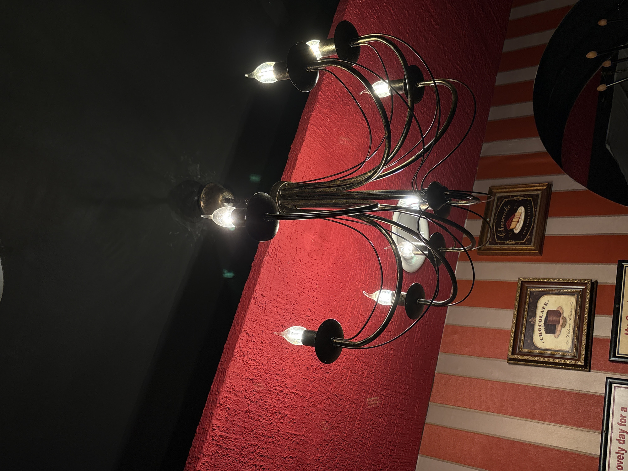The image shows a vintage-style chandelier with multiple arms and candle-shaped bulbs, hanging from a textured red ceiling. The chandelier is dark metal and has a complex, swirling design. Below the red ceiling is a wall with alternating vertical stripes of orange and beige. Two framed pictures hang on the striped wall; one depicts a chocolate cake, and the other shows a slice of cheesecake with the text "Cheesecake New York Style". The scene suggests an indoor establishment, possibly a cafe or restaurant, with a decorative, retro ambiance. The image was captured in Chaikovsky, Russia.