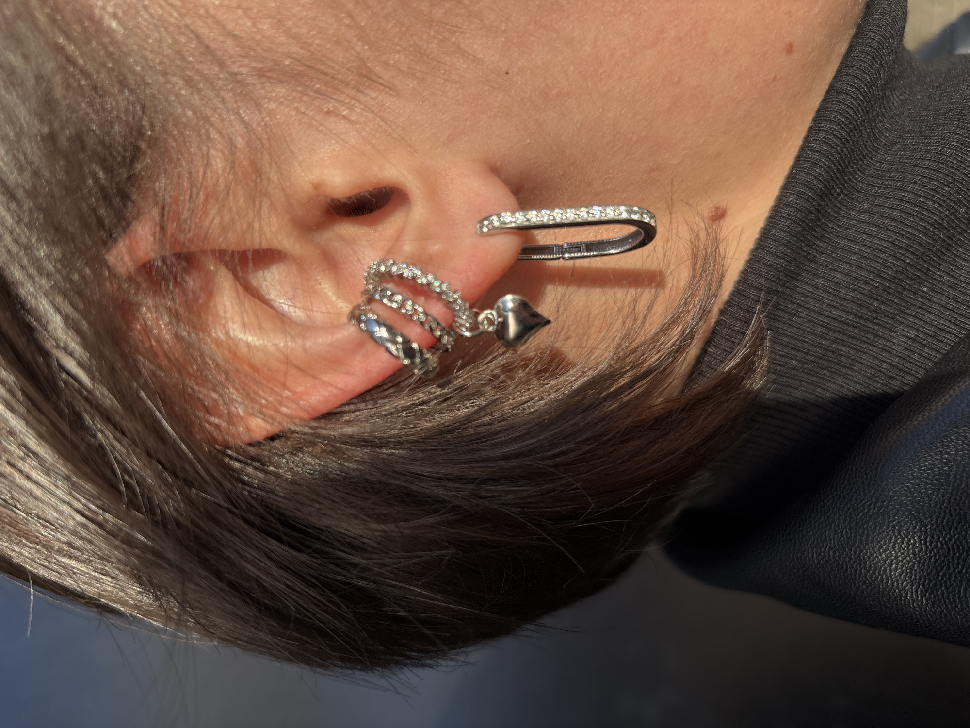 A close-up shot focuses on a person's ear adorned with two distinct pieces of jewelry. The lower piece is a spiral earring with a heart-shaped pendant, while the upper piece is a curved bar studded with stones. The person has short, dark hair. They are wearing a dark garment, possibly leather. The picture is likely taken outdoors in daylight. The location is not identifiable as Izhevsk, Russia from the visual information provided.