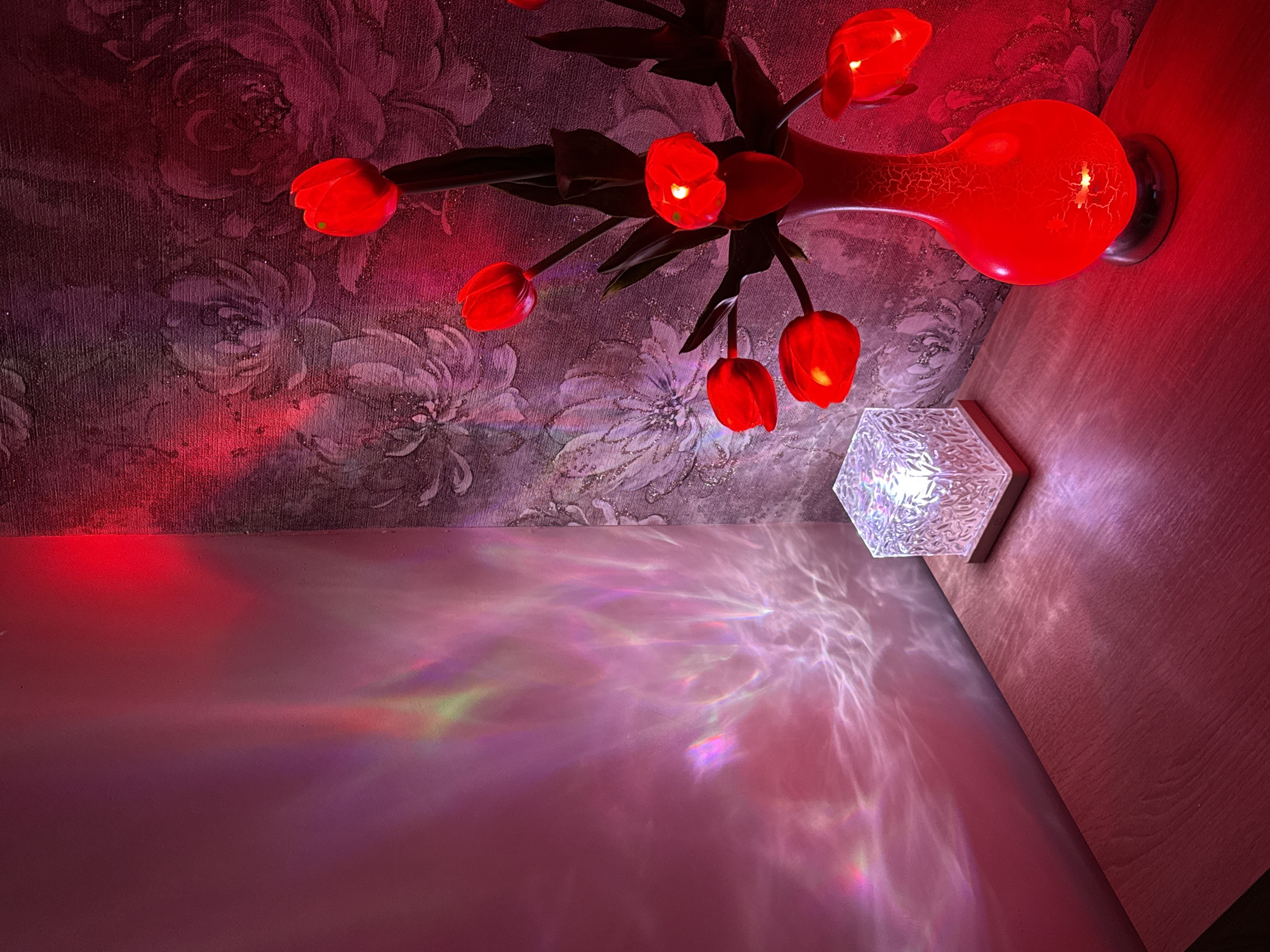 The image presents an interior view, likely taken from a low angle, encompassing a wall and ceiling juncture. The ceiling features floral wallpaper, predominantly purple and pink hues. A red glass fixture resembling stylized tulips is affixed to the ceiling, emitting a red light. Adjacent to the floral fixture, a white geometric light fixture is mounted on the wall, projecting a complex array of light patterns onto the lower wall, casting a rainbow effect. The wall itself appears smooth and painted a light pink. The photograph is brightly lit.