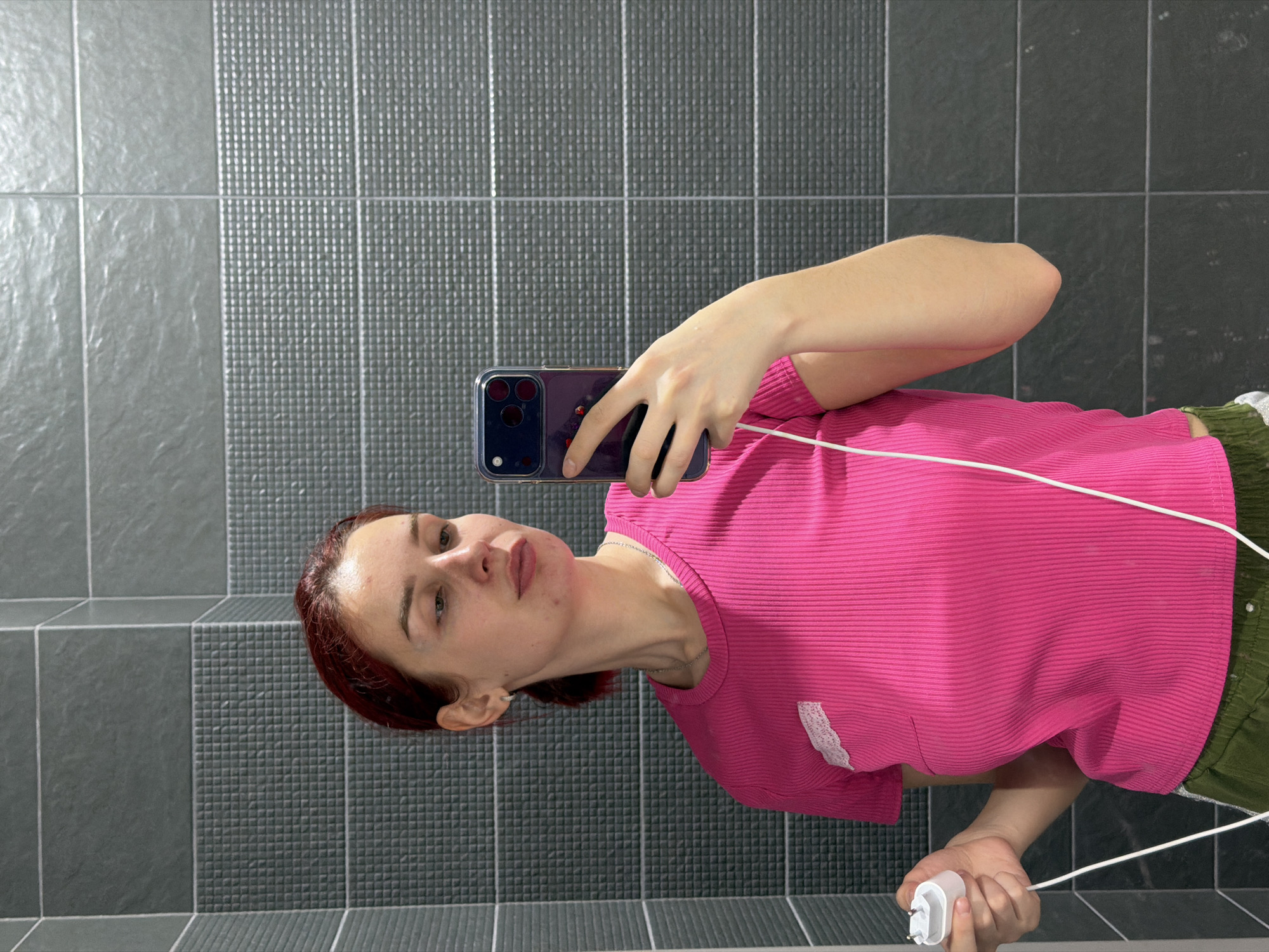 A young woman is taking a selfie in what appears to be a bathroom. The wall behind her is tiled with a mix of grey, textured and smooth square tiles arranged in vertical columns. The woman has reddish-brown hair and is wearing a bright pink, ribbed shirt and green shorts. She is holding a smartphone in her left hand, presumably to take the photo, and a white phone charger adapter in her right hand. The phone has a dark-colored case, and a charging cable is draped across her body. The scene is lit brightly, and the overall impression is of a casual, everyday moment. The location is likely a private residence or a public restroom. There is no indication as to whether the location is in Chaikovsky, Russia, beyond the visual contents of the image.