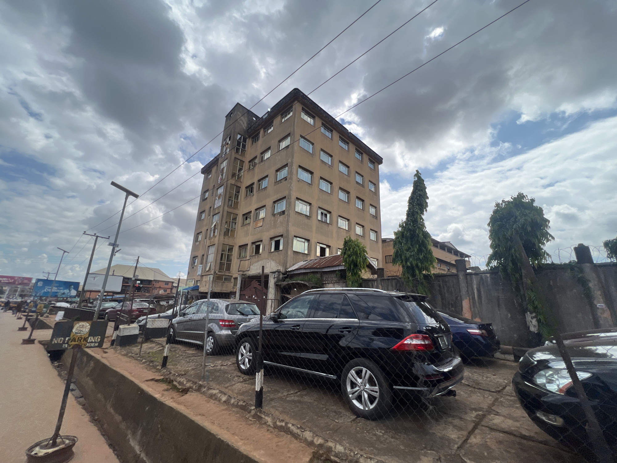 The image depicts a street scene in Nkpor, Nigeria, under a cloudy sky. In the foreground, a black Mercedes-Benz SUV is parked behind a chain-link fence. To its left, a gray Mercedes-Benz sedan is also visible. The vehicles are parked on a paved area adjacent to a raised concrete drainage channel.
Dominating the background is a multi-story, weathered building with a somewhat utilitarian architectural style. It has numerous windows and appears to be an office or residential block. Several utility poles with wires are visible in front of the building, suggesting an urban or semi-urban environment.
Further down the street, more buildings and vehicles can be seen, including what appears to be a billboard. A prominent "NO PARKING BY ORDER" sign is placed on the left side of the image, near the concrete barrier. The overall impression is that of a busy, functional area, possibly a commercial district. The weather appears overcast, with patches of blue sky visible through the clouds. The time of day is difficult to ascertain precisely due to the cloud cover but seems to be daylight hours.