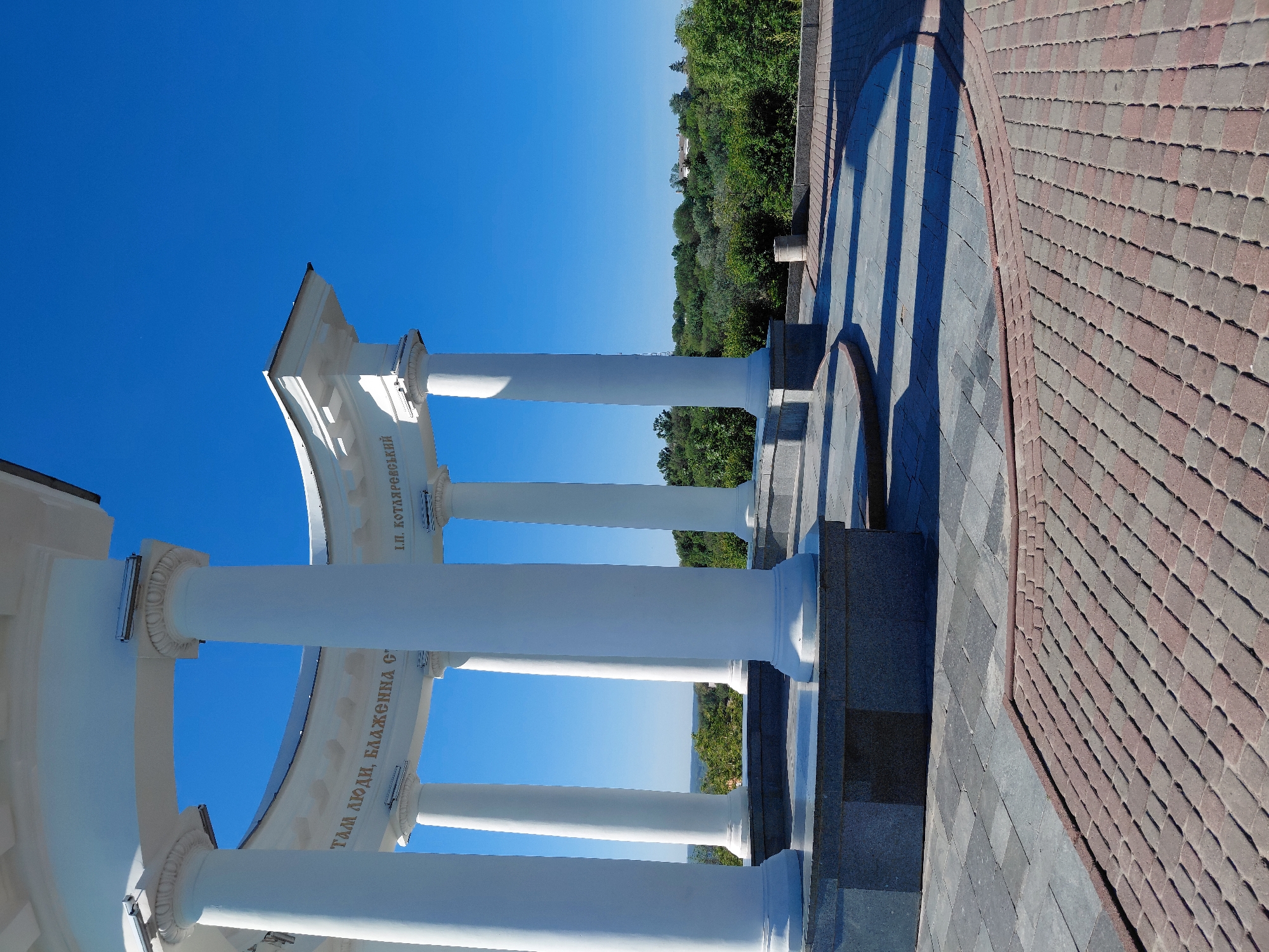 This vertical, brightly lit image captures a section of a classical rotunda or colonnade in Poltava, Ukraine, seen from a slightly low angle looking upwards and outwards. The main subjects are several pristine white, smooth columns supporting a curved entablature. Beyond the columns, a vibrant blue, cloudless sky stretches above a distant horizon line dotted with lush green trees and glimpses of a settlement on a hillside.
The entablature bears inscriptions in golden Cyrillic text. Clearly visible are "І.П. КОТЛЯРЕВСЬКИЙ" (I.P. Kotliarevskyi), referring to the famous Ukrainian writer, and partially visible "ТАМ ЛЮДИ, БЛАЖЕННА С" (There people, blessed c...), which is likely part of a known quote from his works, such as "щасливі там люди, блаженна сторона" (happy there people, blessed country).
The ground in the foreground is paved, featuring two distinct patterns: grey square pavers closer to the structure and reddish-brown rectangular pavers forming a path or plaza further out. Strong, elongated shadows cast by the columns fall across the paving, indicating bright sunlight and suggesting either morning or late afternoon. The weather is clear and sunny. No people are visible, allowing the architectural grandeur and the serene natural surroundings to dominate the scene.