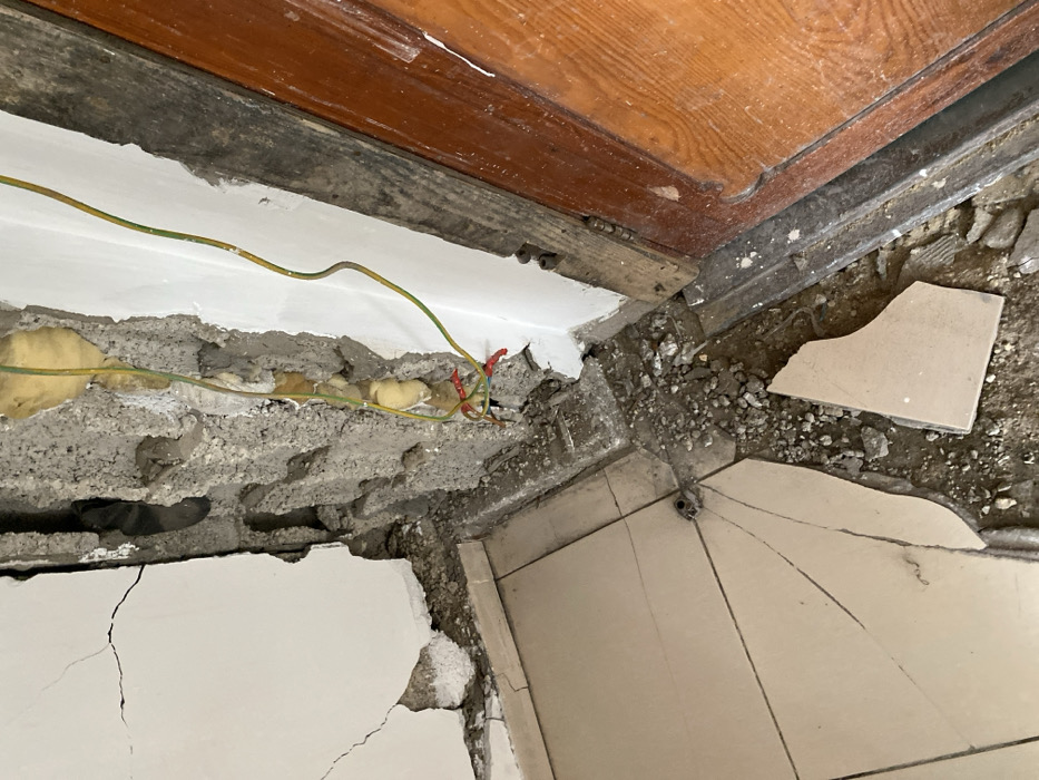 The image depicts severe damage within an interior corner of a structure in Khan Yunis, Palestinian Territory. The scene is characterized by a fragmented white plastered wall on the left, showing multiple large cracks and missing sections of plaster, exposing underlying grey material. Adjoining it, the right wall is significantly compromised, with extensive portions of concrete or masonry removed, revealing rough grey internal structure and some yellow, fibrous insulation-like material. Two electrical wires, one yellow with a green stripe and another green with a yellow stripe, hang loose from the damaged right wall, with their exposed ends showing red and yellow insulation from smaller connected wires. The floor is tiled with light-colored, possibly beige, square tiles, many of which are broken, cracked, or completely dislodged, exposing dirt and debris underneath. A substantial amount of grey rubble, dust, and fragments of concrete and plaster are scattered across the floor, concentrated at the base of the damaged walls and around the corner. To the far right, a section of a dark brown wooden door or door frame is visible, also surrounded by debris at its base. No people are present, and no active events are depicted; the scene represents the aftermath of structural damage.