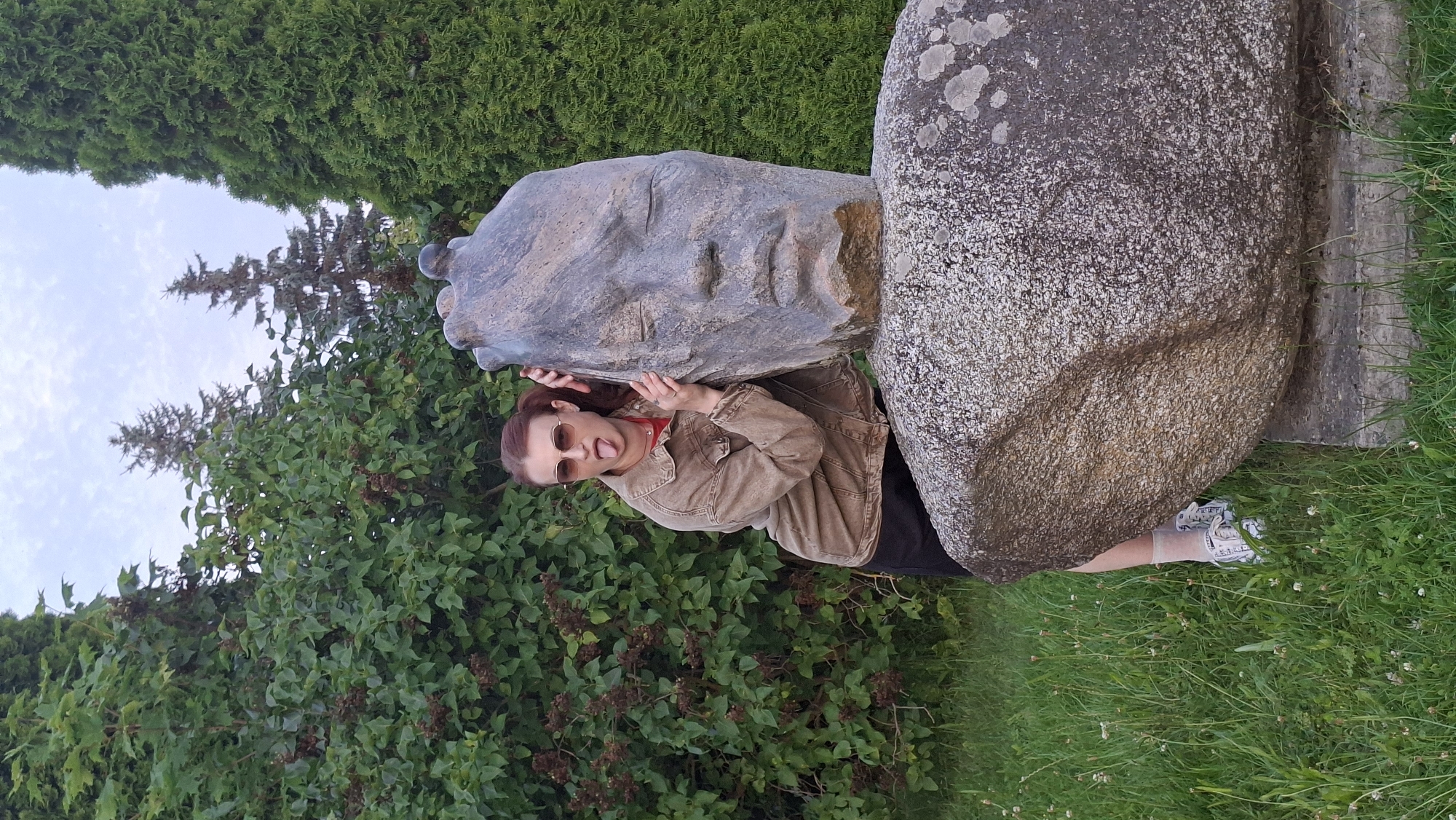 In the city of Myadzyel, Belarus, a woman with auburn hair pulled back in a ponytail, wearing sunglasses and a beige jacket over a red shirt and black shorts, is playfully leaning against a large stone sculpture of a human head. Her tongue is sticking out as she rests her hands on the sculpture. The sculpture, weathered and grey, appears to be part of a larger stone structure, possibly a monument, set against a backdrop of lush green trees and foliage. The ground is covered in green grass with scattered small white flowers. The sky above is overcast with light clouds.