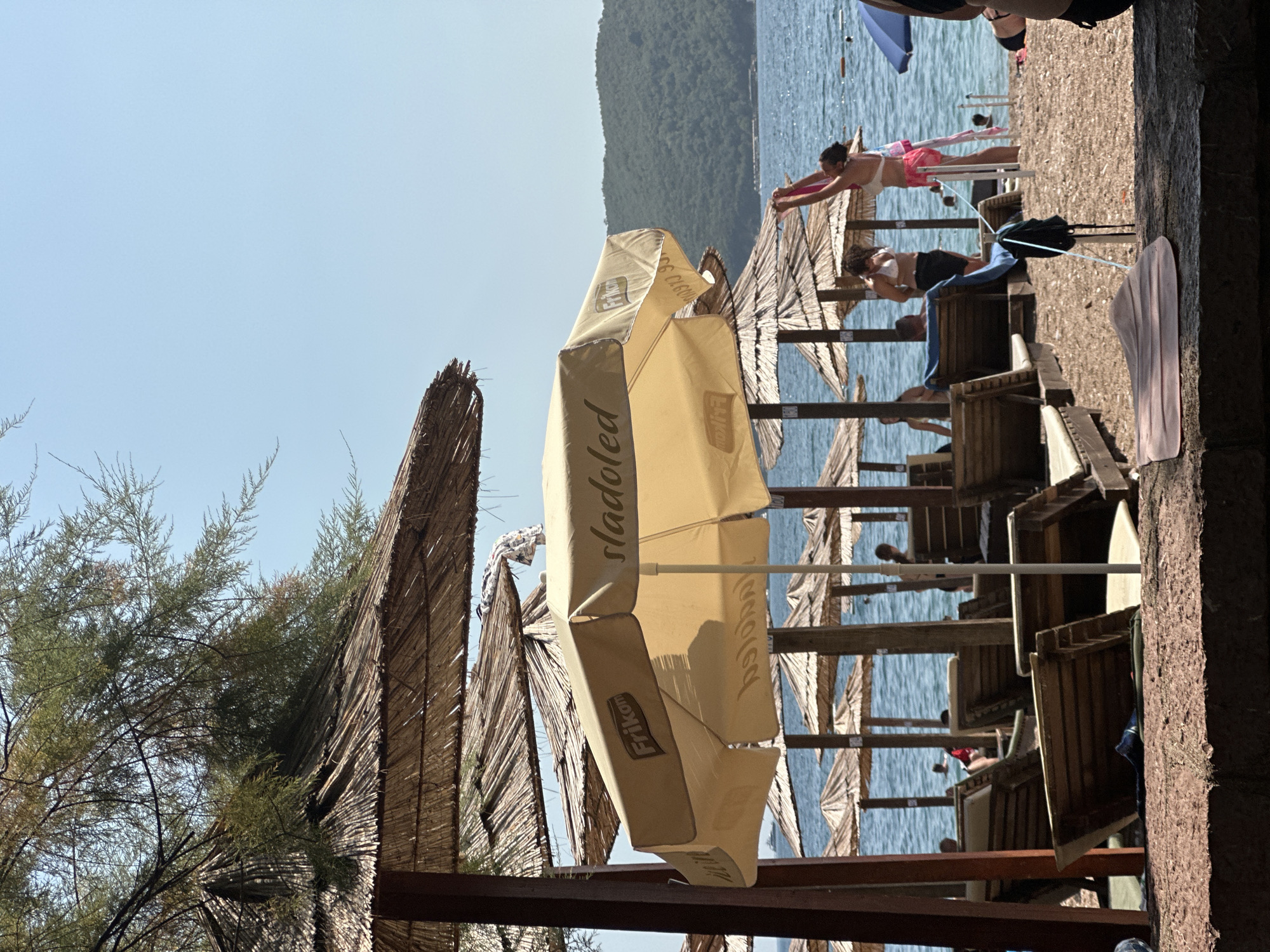 The image shows a crowded beach scene in Budva, Montenegro, on a sunny day. The foreground is dominated by a light yellow beach umbrella with the word "sladoled" (ice cream in Serbian/Croatian) and the logo "Frikom" printed on it. Several other straw-covered umbrellas are visible in the background, providing shade for rows of empty wooden lounge chairs.
The beach is made of pebbles and sand. In the middle ground, several people are enjoying the beach. A woman in a white bikini and pink shorts is standing and stretching her arms upwards. Another woman in a white bikini and dark shorts stands nearby. Other beachgoers are seen in the water and scattered around the beach, some under umbrellas. The blue sea stretches to the horizon, with a green, tree-covered hill forming the backdrop. The sky is clear and blue. The overall atmosphere is that of a typical summer day at a popular tourist destination.