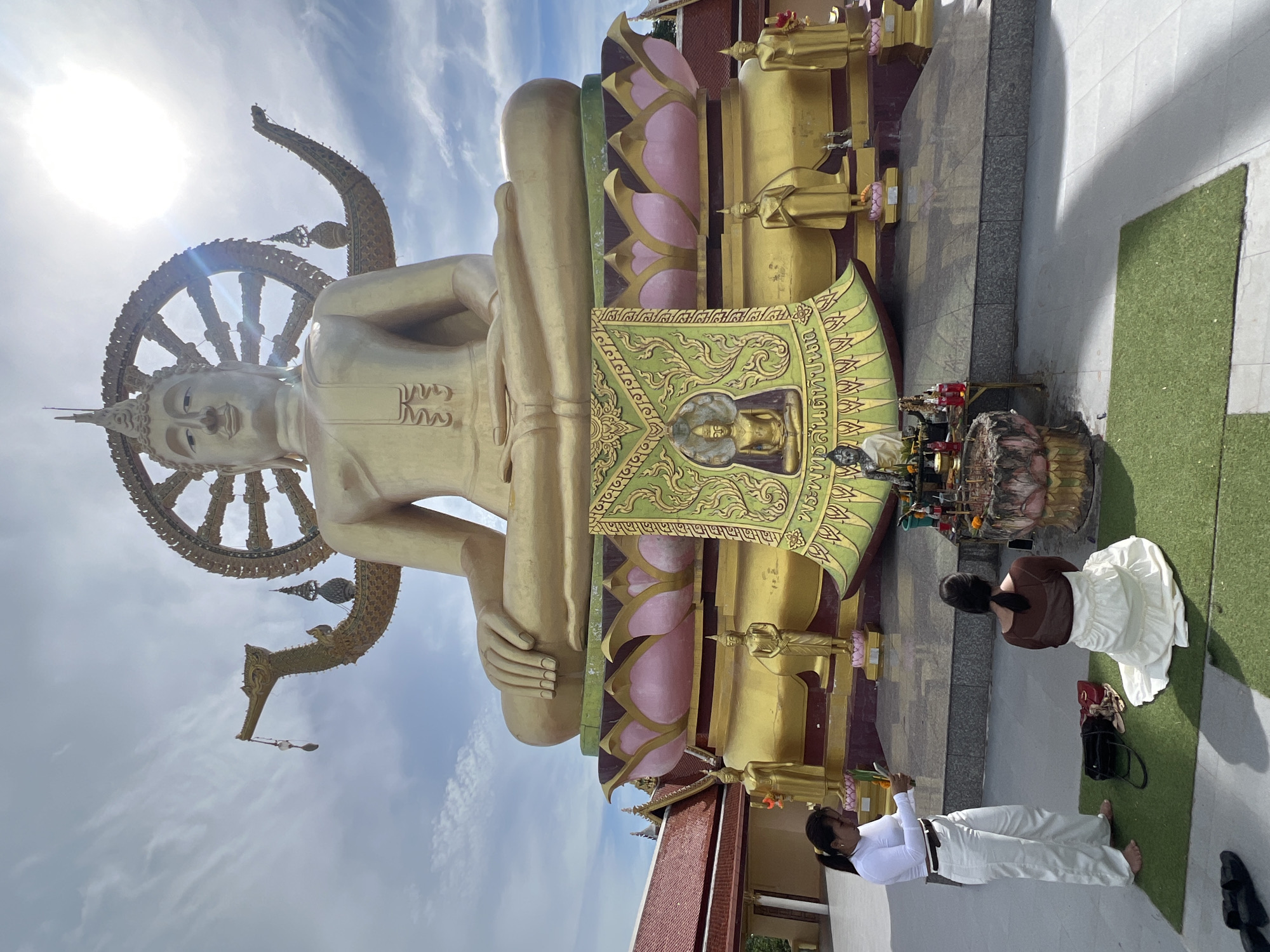 The image shows the Big Buddha of Koh Samui, located at Wat Plai Laem temple in Baan Plai Laem, Thailand. A large, golden statue of Buddha is seated in a meditative pose, with a large ceremonial wheel behind it, against a partly cloudy sky illuminated by the sun. In the foreground, two women are present. One woman, wearing a white top and white pants, stands on a tiled surface and holds what appears to be offerings. Another woman, dressed in a white skirt and a brown sheer top, kneels on a green mat in front of an altar, which contains incense sticks, candles, and offerings. Several small golden statues of Buddha and other figures are placed around the base of the main statue. The environment is outdoors, with a tiled walkway and a glimpse of a temple building with a red roof to the left.