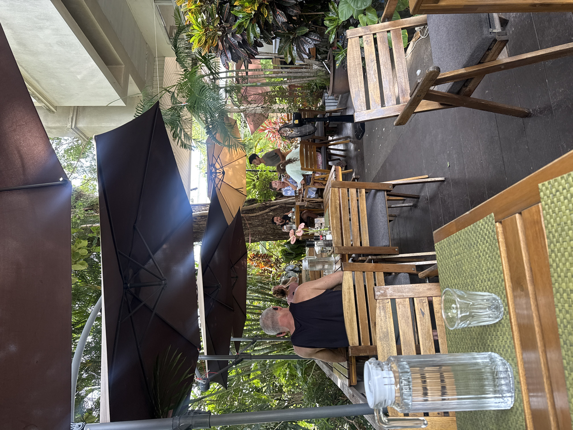The image depicts an outdoor dining area at a restaurant in Baan Chaweng Noi, Thailand. Several wooden tables and chairs are arranged on a dark wooden deck. Large, dark brown umbrellas are providing shade. Lush green foliage and palm trees surround the dining area.
In the foreground, a glass pitcher filled with water and a small drinking glass sit on a table covered with a green placemat. A man wearing a black tank top is seated at a table in the mid-ground, facing away from the camera. Several other patrons are visible at various tables in the background, some engaged in conversation. A ceiling fan is positioned between tables in the mid-ground. The scene is brightly lit, suggesting it is daytime.