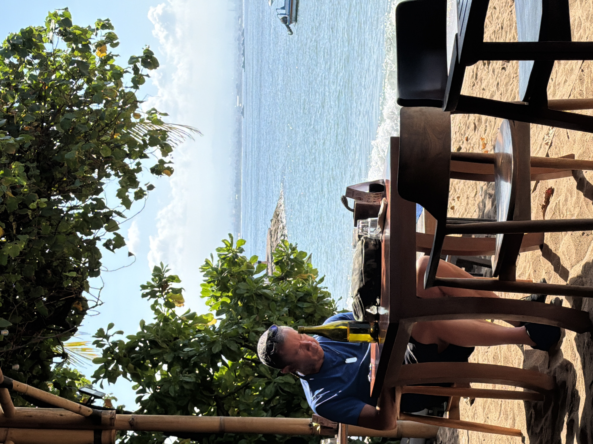 A man wearing a blue t-shirt and black shorts sits at a wooden table on a sandy beach, looking down at a bottle of wine. He has sunglasses on his head and a necklace. The table is set with a bottle of wine, a bag, and glasses. In the background, the ocean is visible with a boat in the distance. The scene is lit by bright sunlight, casting shadows on the sand. The man appears to be at a beachside cafe in Denpasar, Indonesia, possibly Sanur beach, known for its relaxed atmosphere and ocean views.