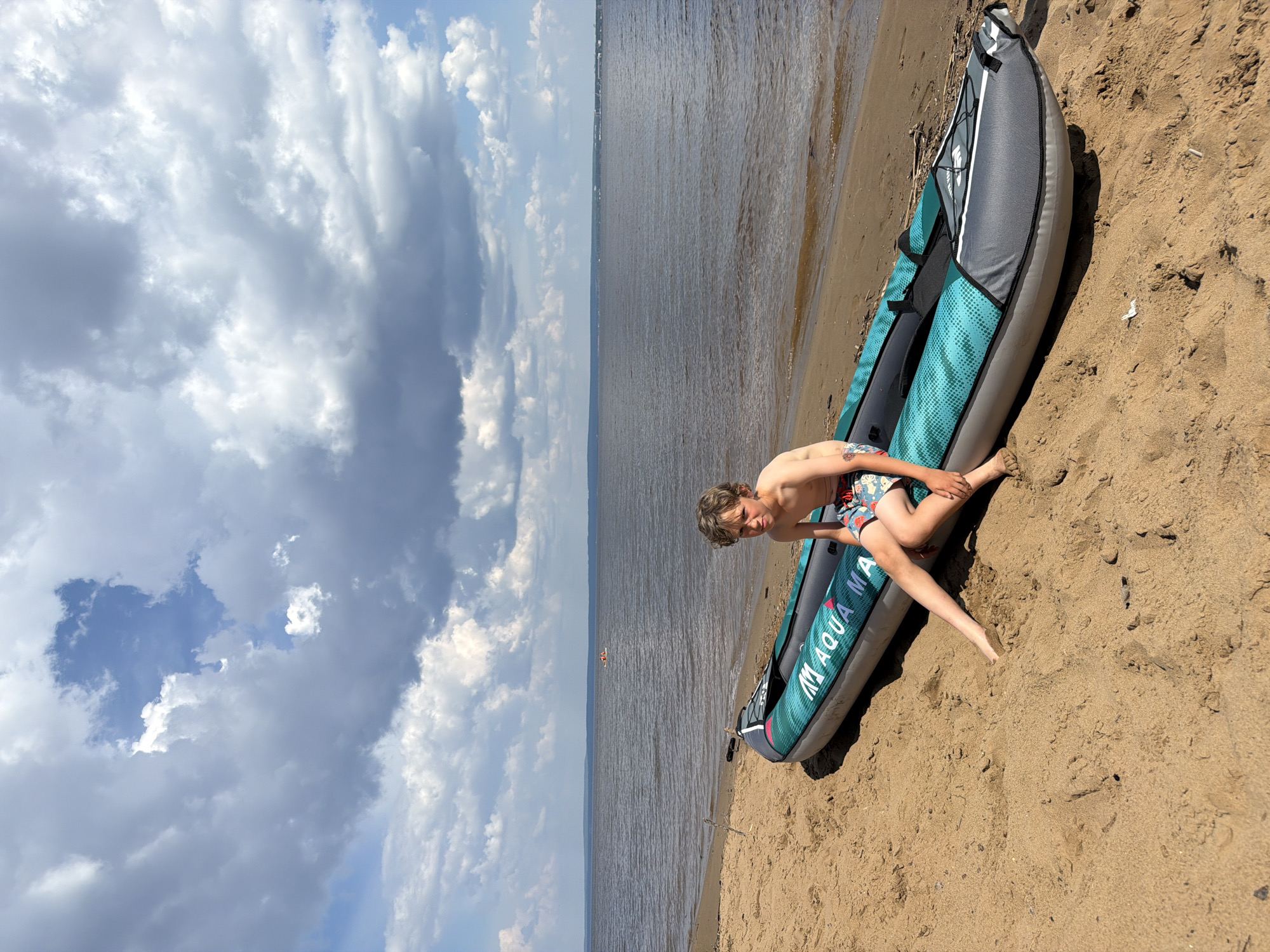 A young boy with curly brown hair and a serious expression sits on the edge of a deflated, gray and teal inflatable kayak on a sandy beach. He is wearing patterned swim trunks. The kayak has "Aqua Marina" printed on its side. Behind the kayak, a calm, wide body of water stretches to the horizon, where a faint line of trees and buildings can be seen. The sky above is a mix of blue and large, dramatic cumulus clouds. In the distance on the water, a small boat with several people is visible. The environment appears to be a natural, outdoor setting, possibly a lake or a wide river, under a partly cloudy sky. The image is taken from a medium-low angle, looking slightly up at the boy and the sky.