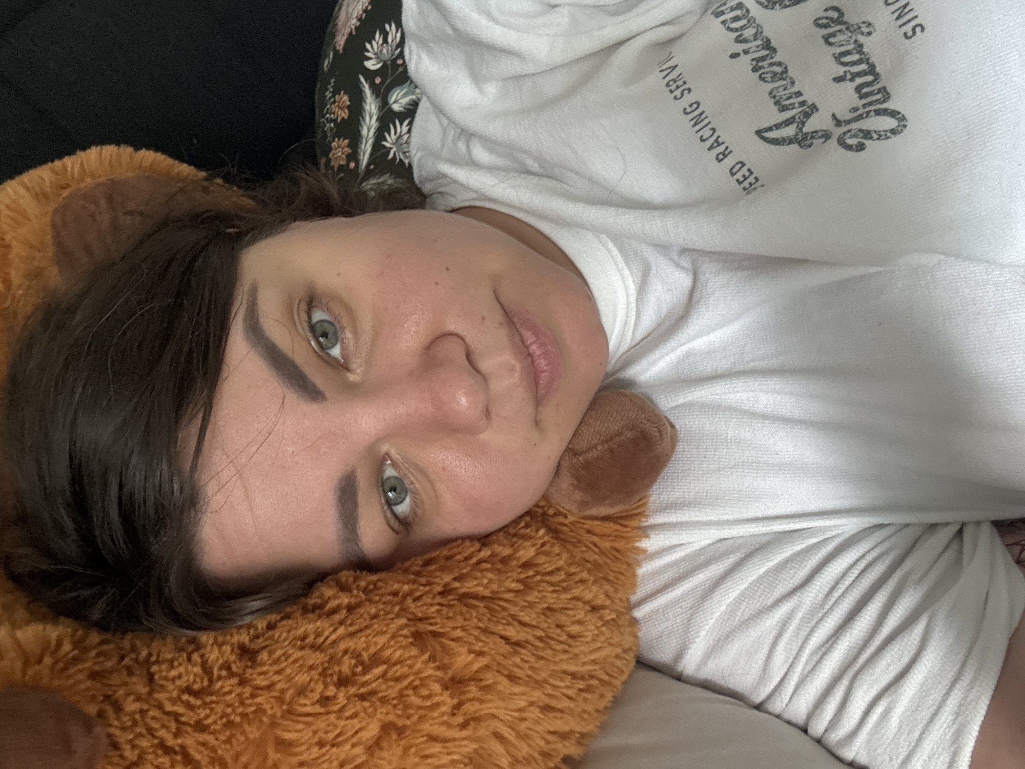 A close-up, overhead selfie captures a woman with brown hair and green eyes lying down, resting her head on a large, fuzzy, brown teddy bear. She is wearing a white t-shirt with a graphic print that reads "SPEED RACING SERVICE American Vintage SINCE". Her skin is flushed, particularly on her cheeks, and she has a neutral expression. The background shows a dark surface and a pillow with a floral pattern. The image appears to be taken indoors. There is no information to determine the location of this image within Höhenkirchen-Siegertsbrunn, Germany.